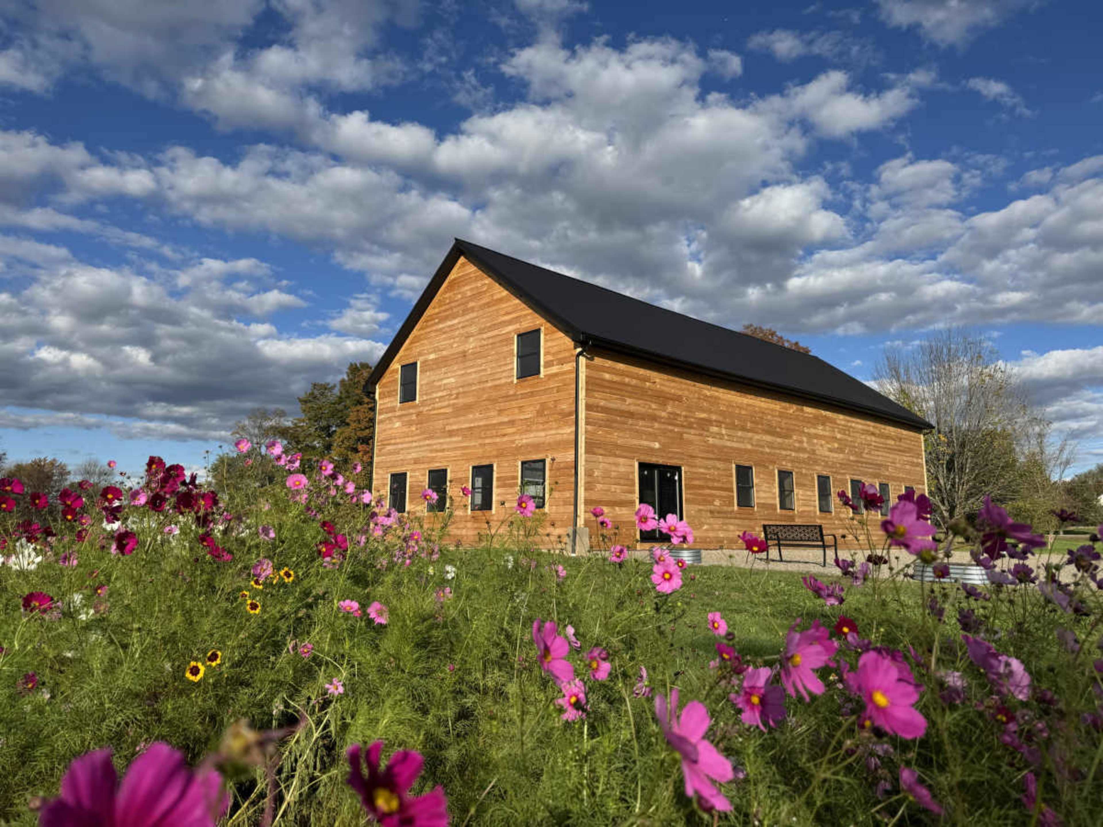 A wooden barn with a black roof stands surrounded by vibrant flowers under a cloudy blue sky.