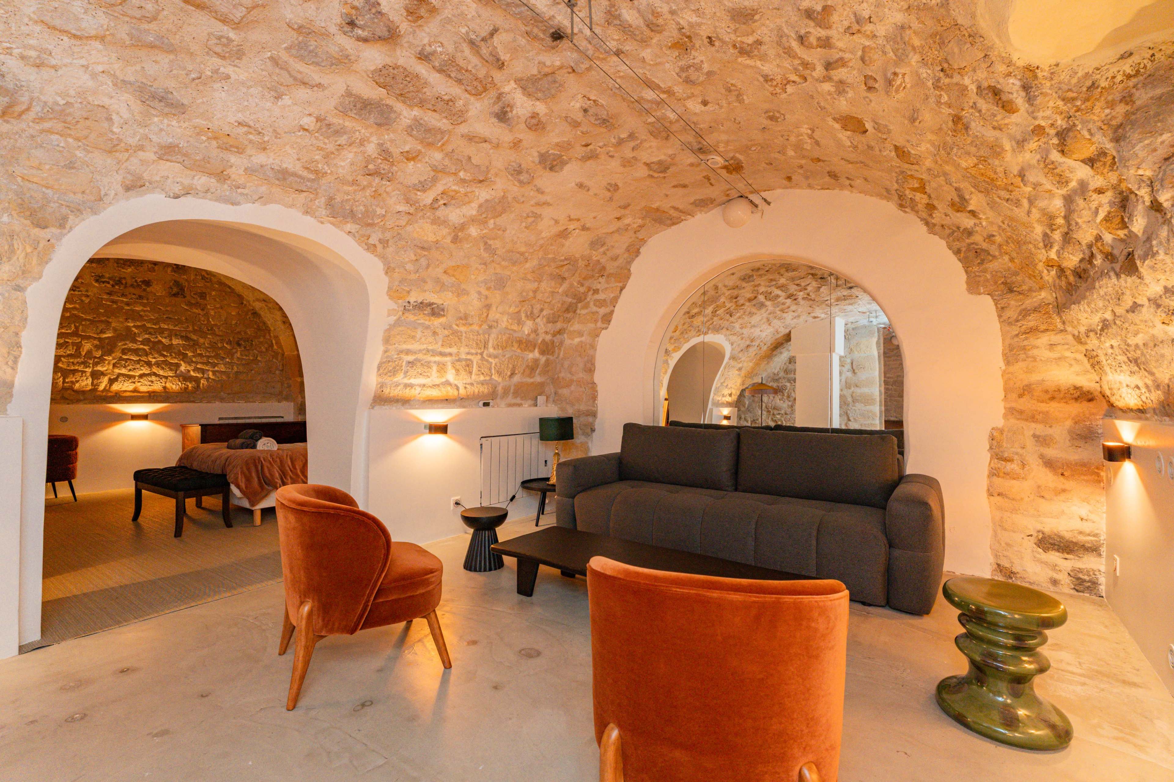 The scene features a stone-walled room with a gray sofa, two orange chairs, and a small table, creating a cozy living space that connects to a bedroom area through arched openings.