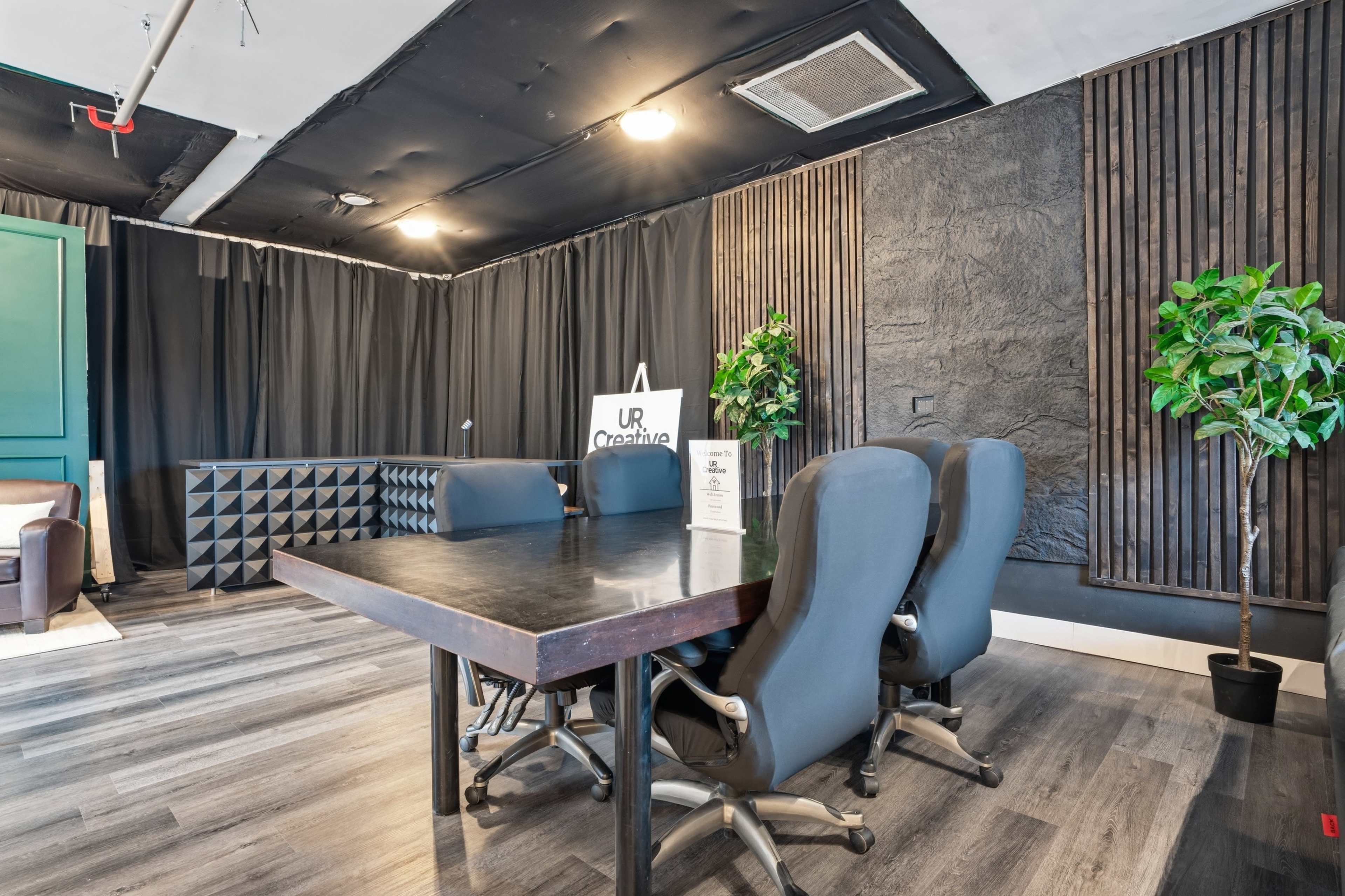 The image shows a modern conference room with a dark table surrounded by ergonomic chairs, accented by textured walls and indoor plants.