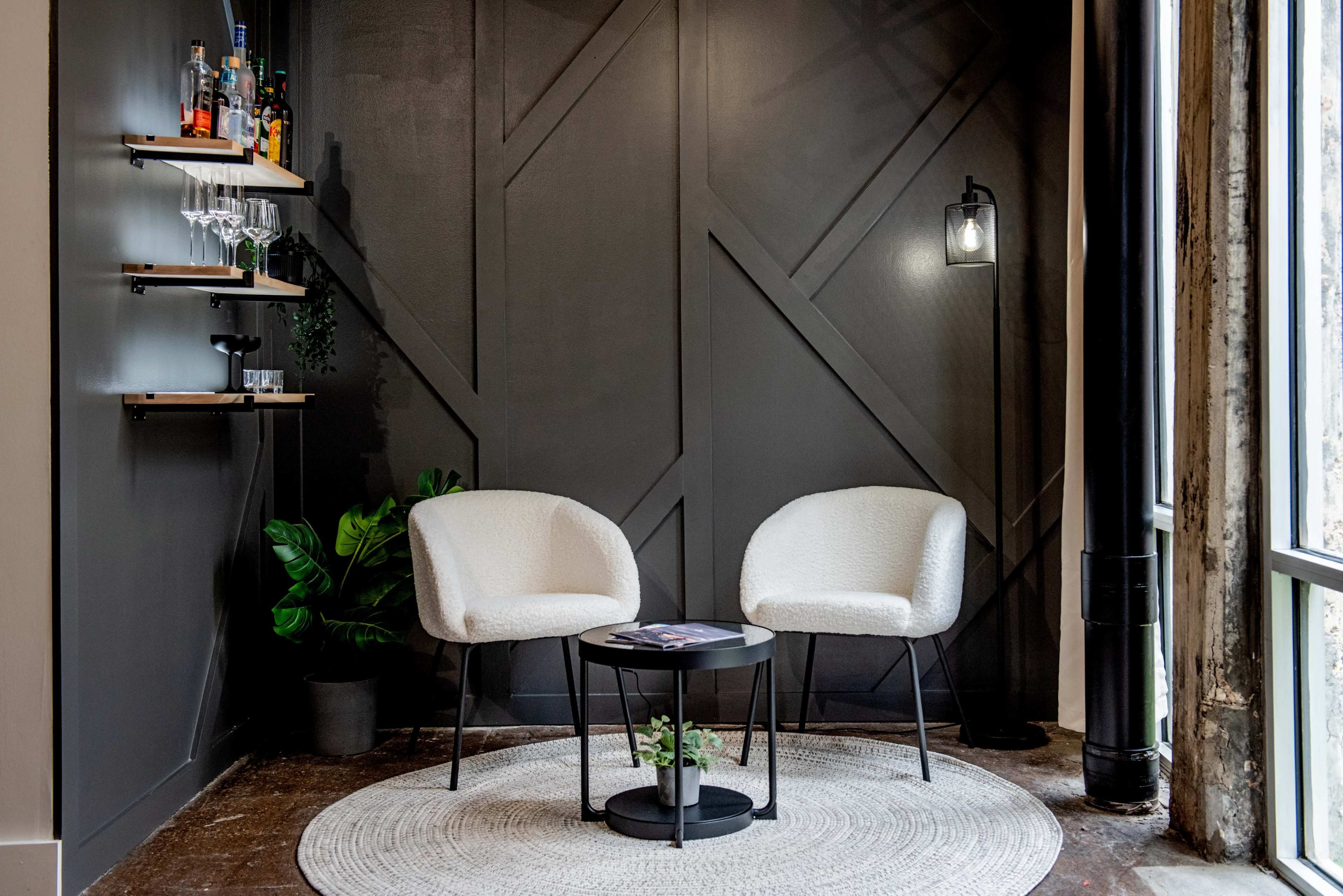 A modern seating area featuring two white chairs, a small black table, and a wall-mounted shelf with drinks and glassware against a dark geometric background.