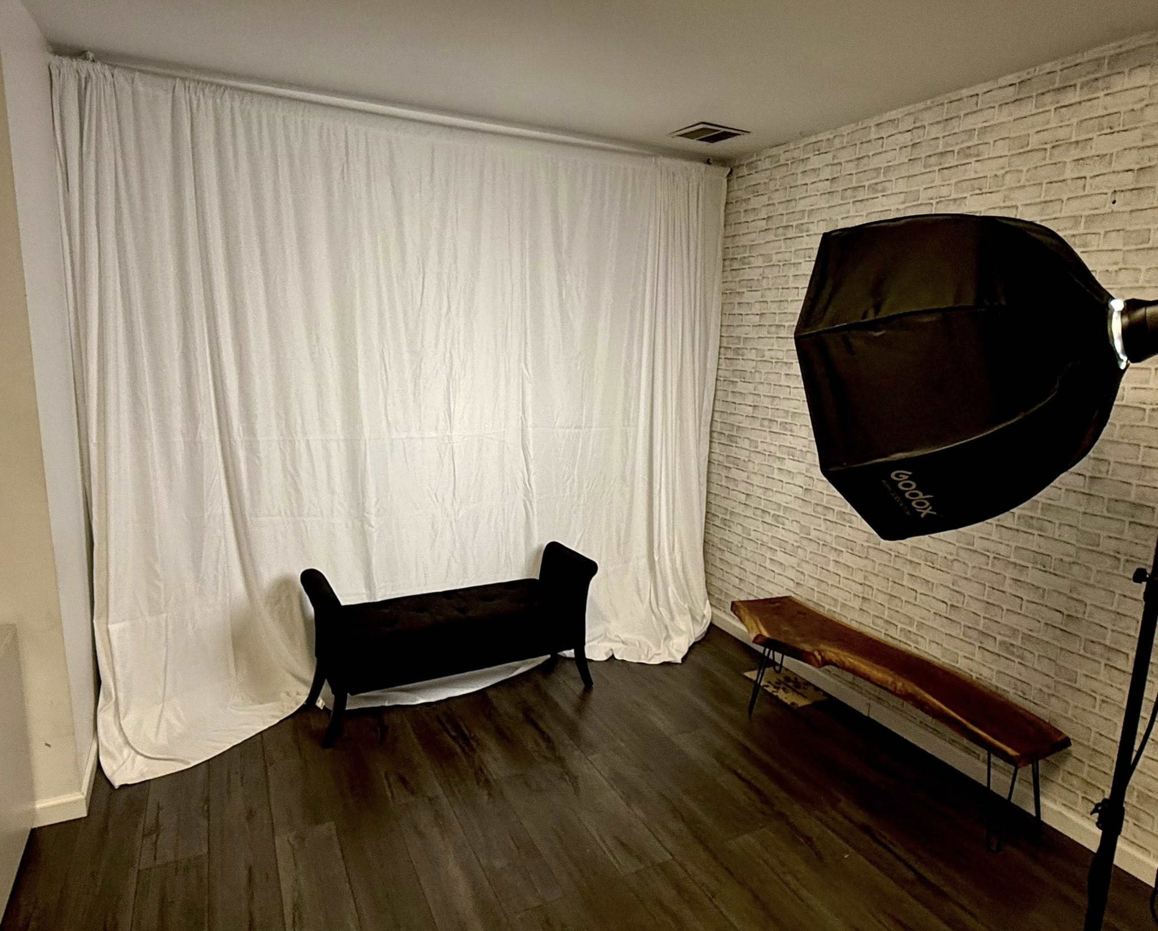 The image shows a photography setup featuring a black couch in front of a white backdrop, with a softbox light and a wooden bench nearby on a wooden floor.