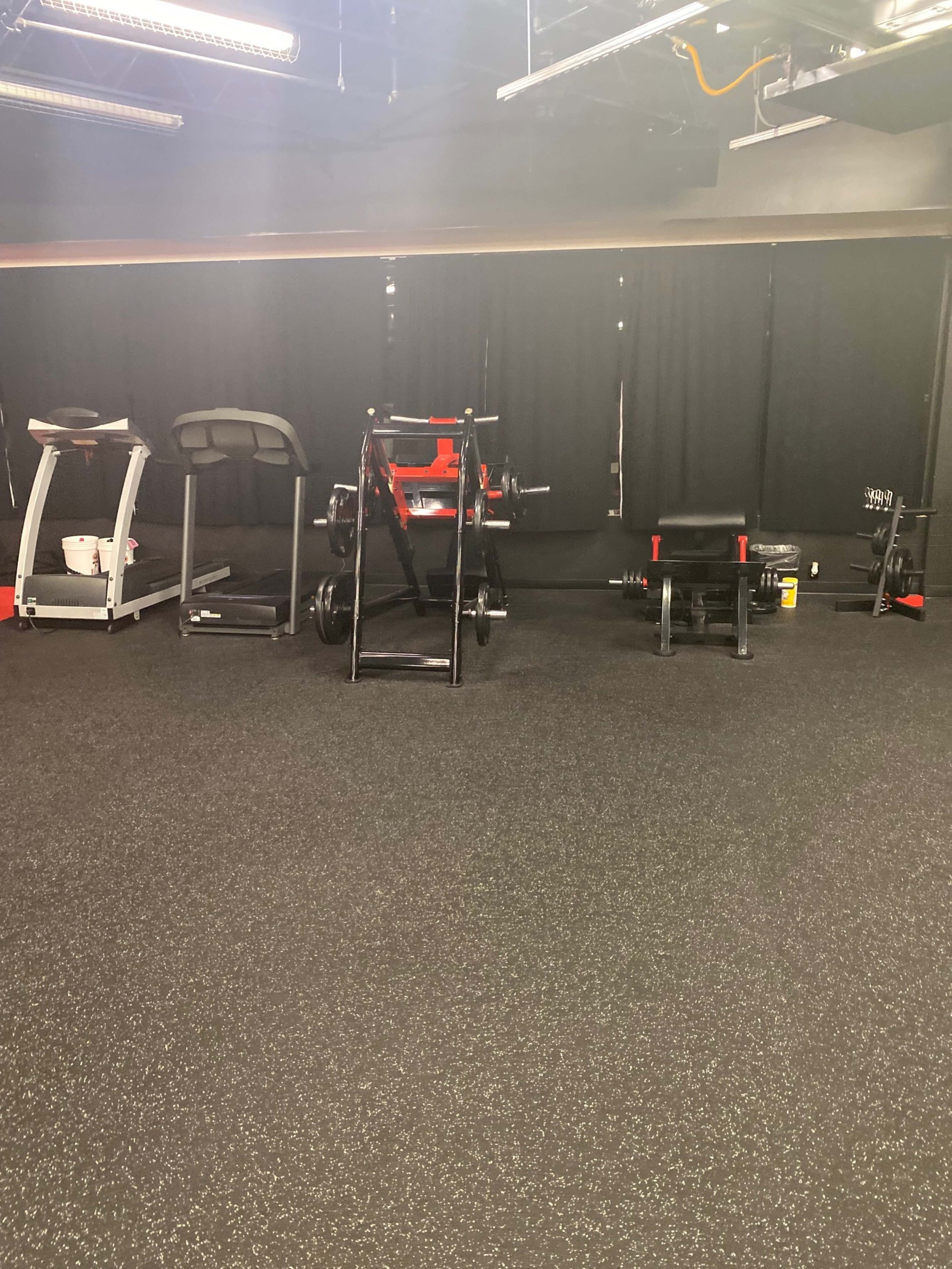 A gym area with various exercise equipment, including a treadmill, weights, and a bench, against a backdrop of black curtains.