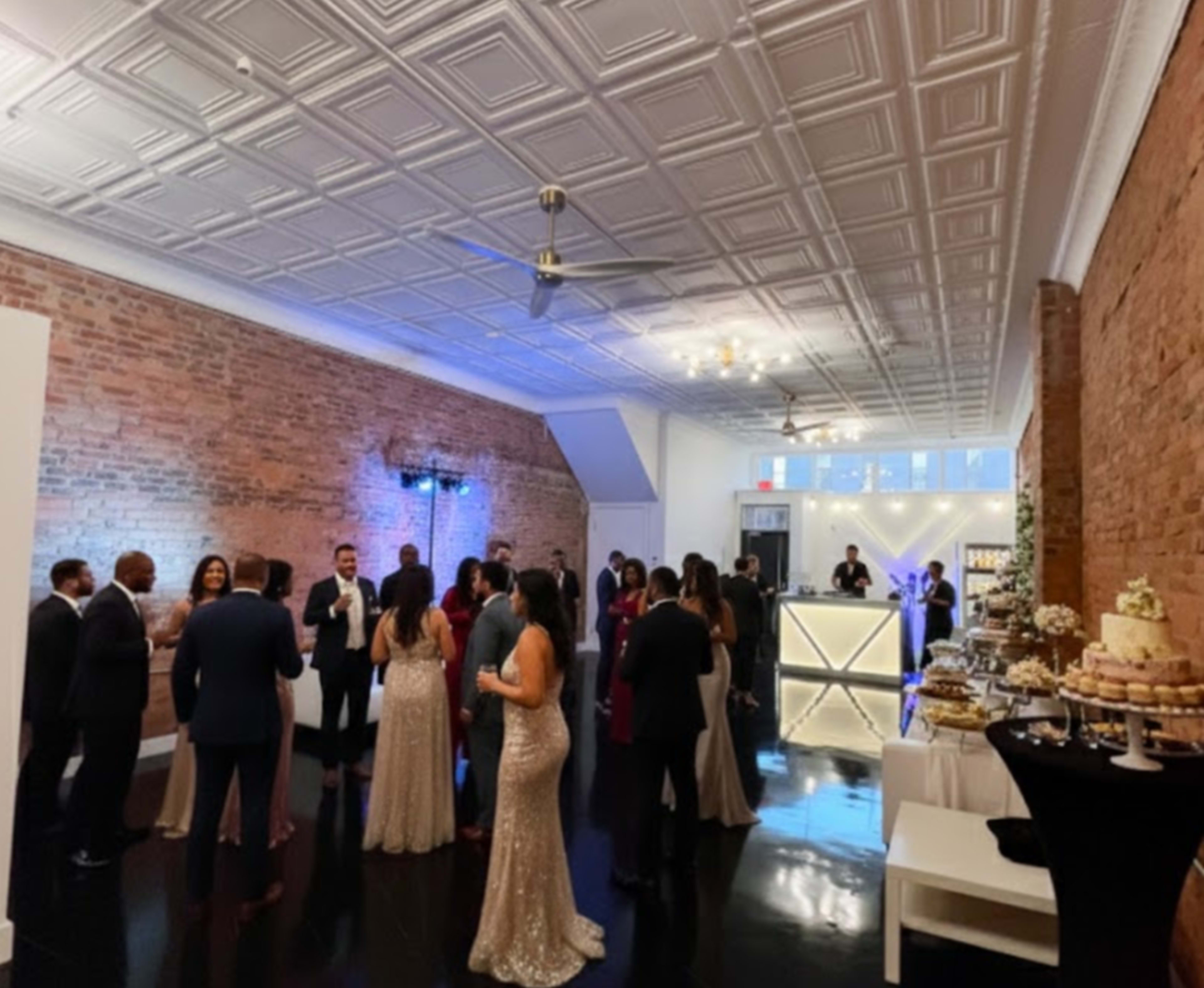 A group of guests in formal attire socialize in a decorated event space with exposed brick walls and a dessert table nearby.