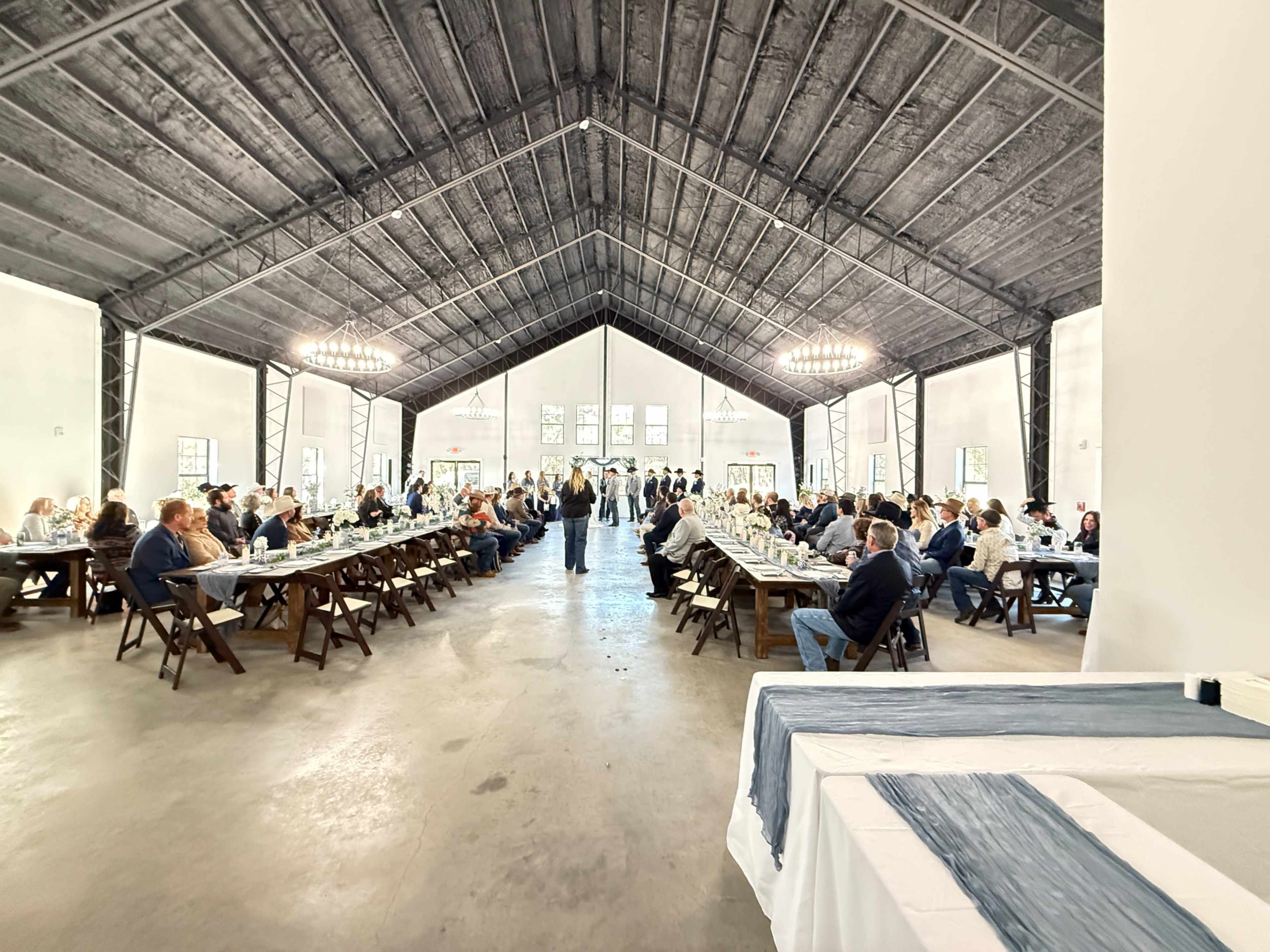 A large group of people is seated at tables in a spacious venue, with a high ceiling and large windows, engaging in an event or gathering.