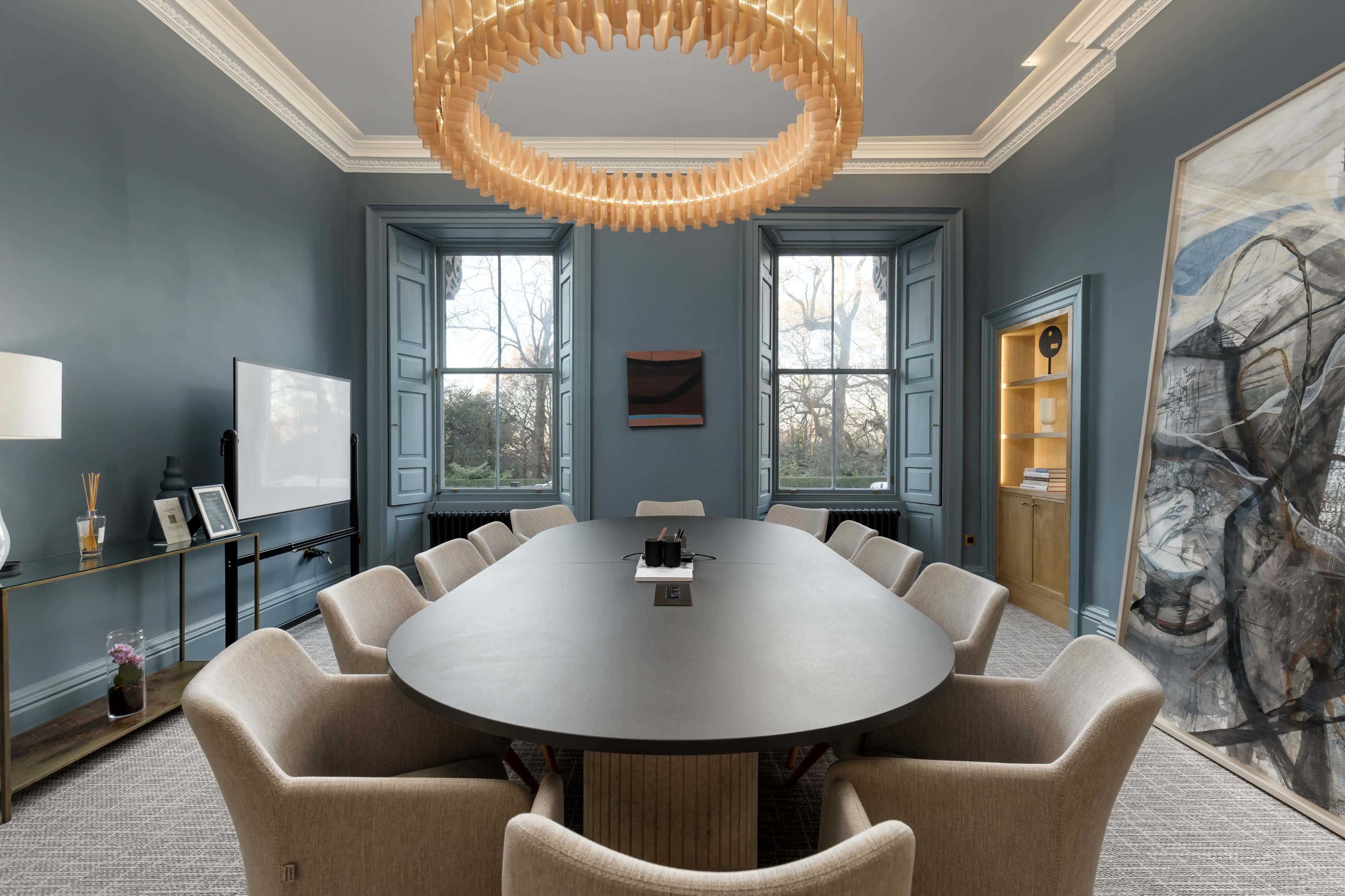 The image shows a spacious conference room with a large oval table surrounded by upholstered chairs, illuminated by a unique chandelier, and featuring tall windows with a view of outdoor trees.