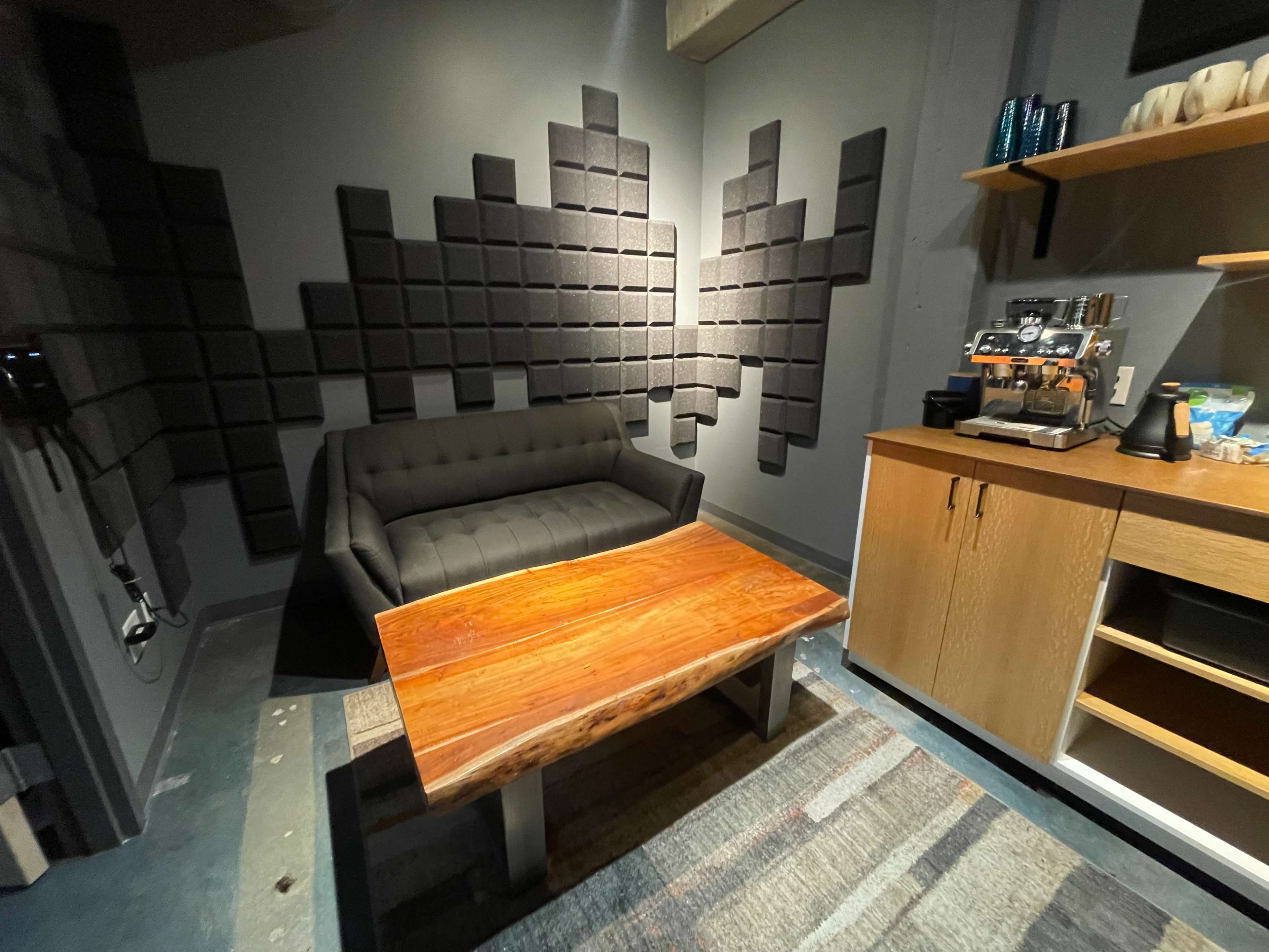 A small room with a black sofa, a wooden coffee table, acoustic paneling on the walls, and a coffee machine on a countertop.