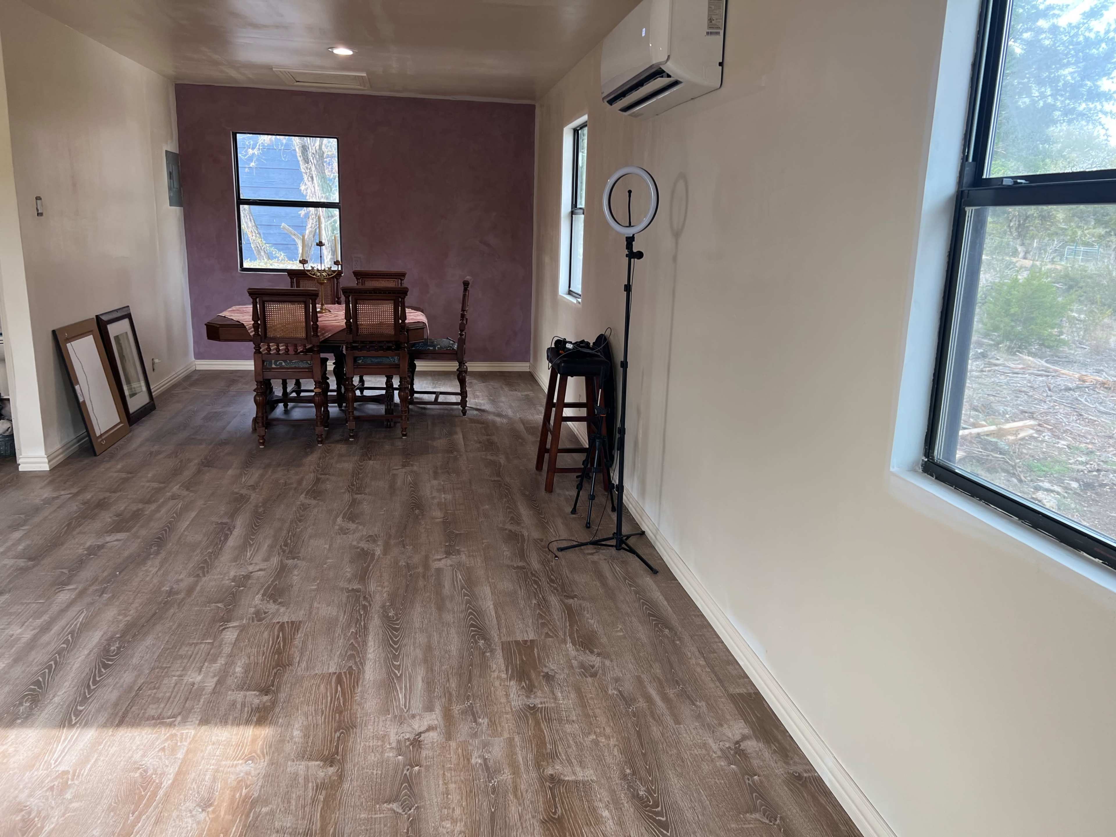 The image shows a well-lit room with wooden flooring, a dining table surrounded by chairs, and a small stand with a ring light in one corner.
