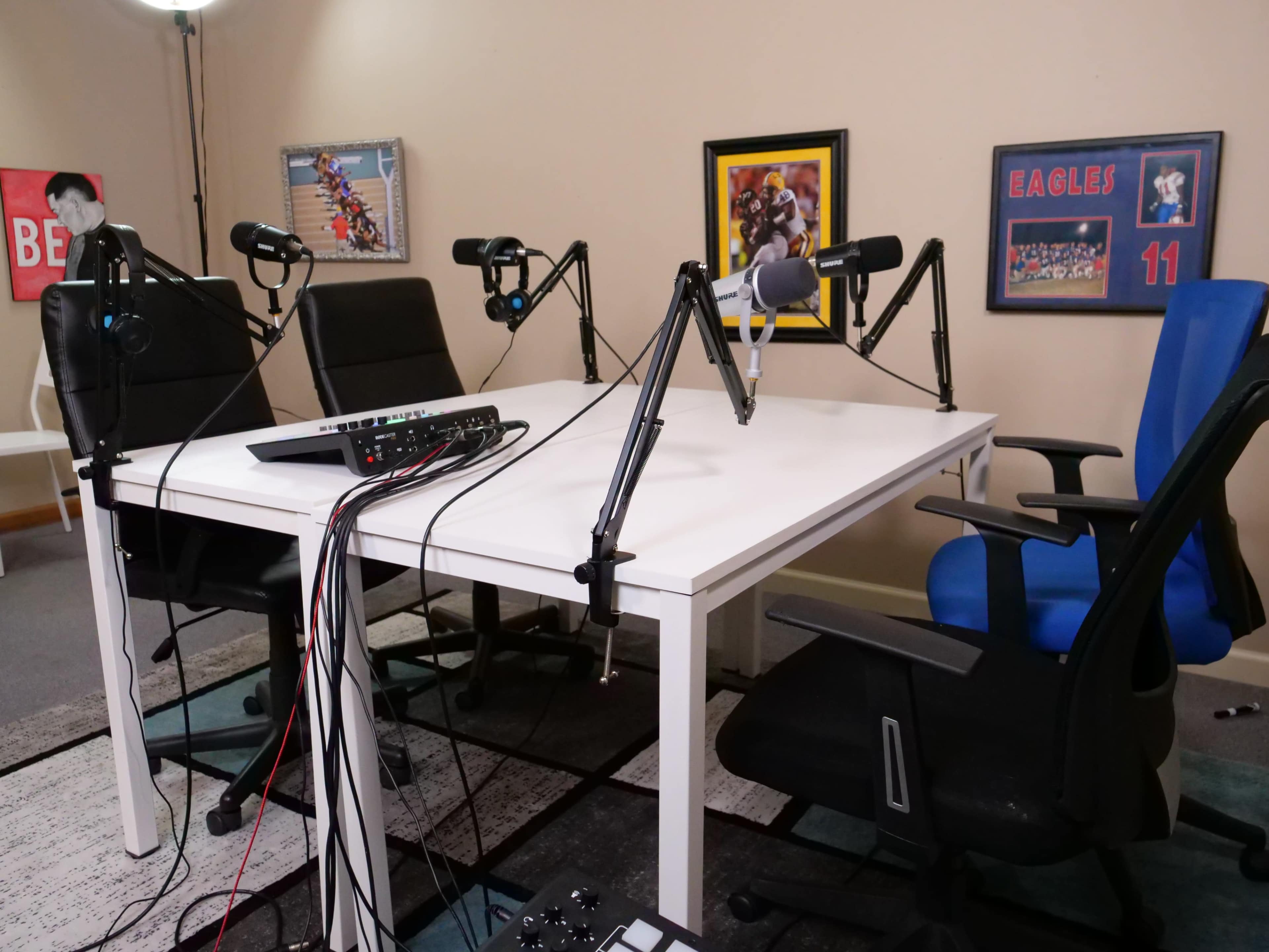 A podcast recording setup with a white table, multiple microphones on stands, and four chairs of varying colors arranged around the table.