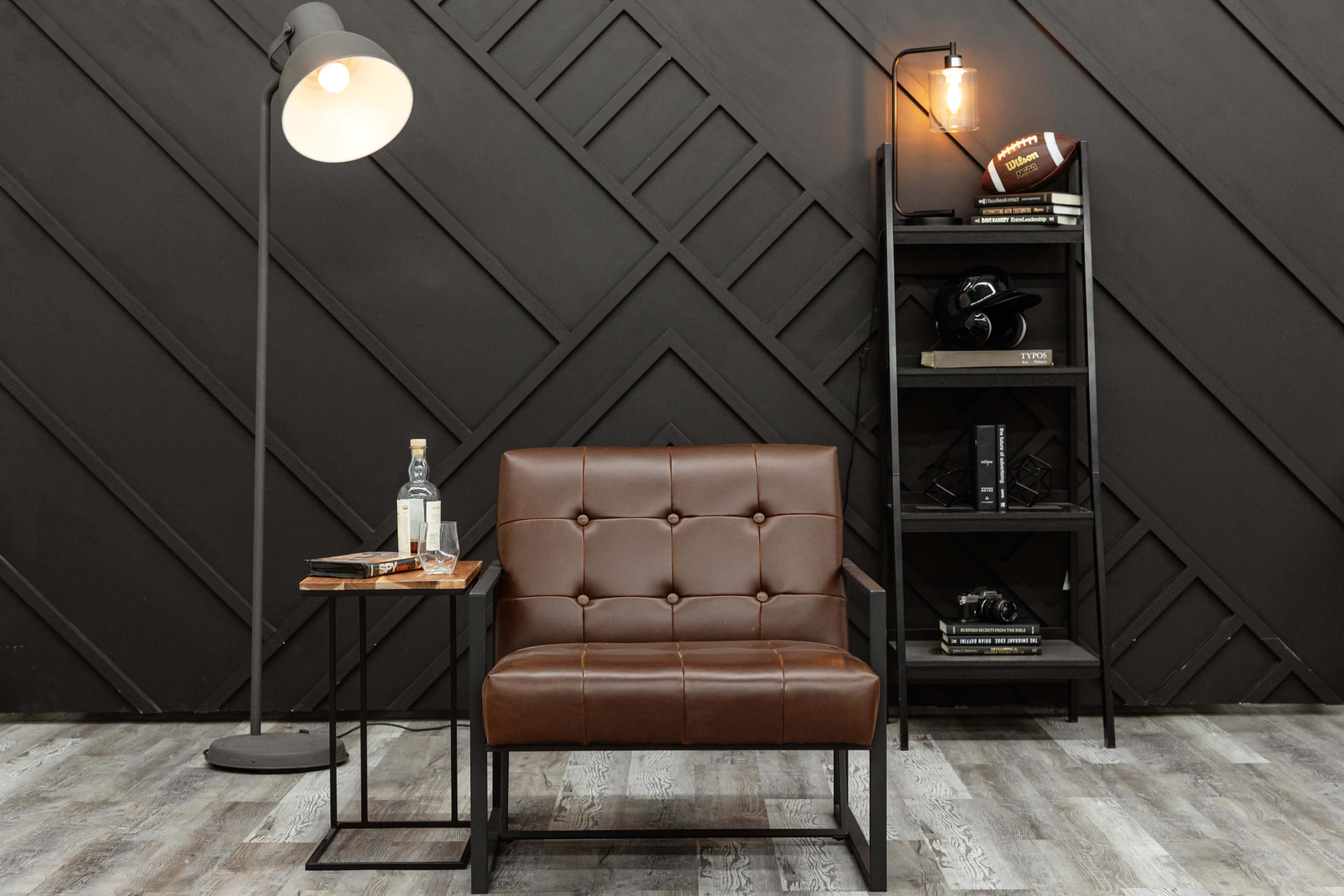 The image shows a modern sitting area featuring a brown leather chair beside a small table with a bottle and glass, a floor lamp, and a black bookshelf displaying books and decor against a textured dark wall.