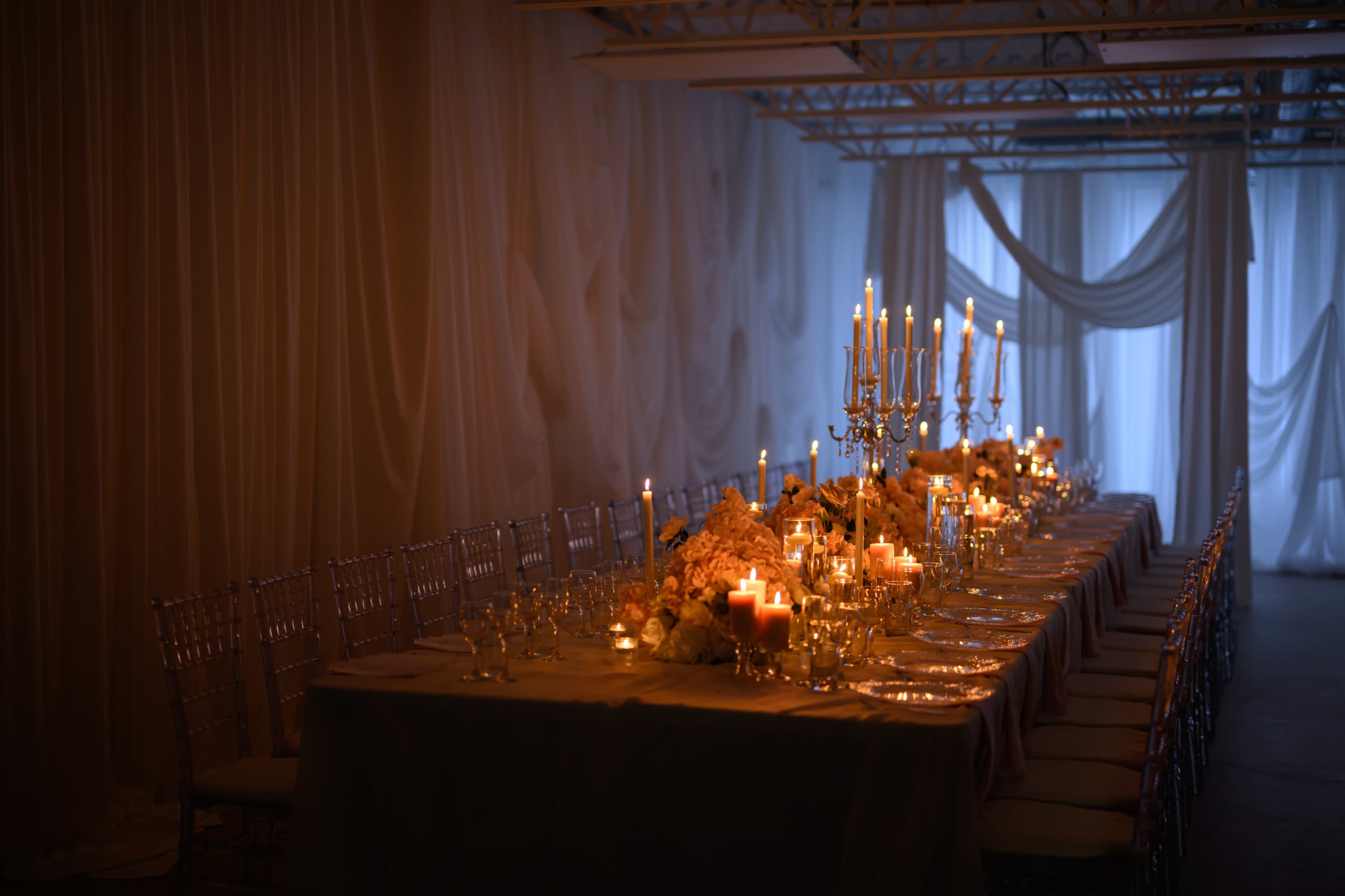 A long, elegantly set dining table is adorned with candles and floral arrangements in a softly lit, draped room.