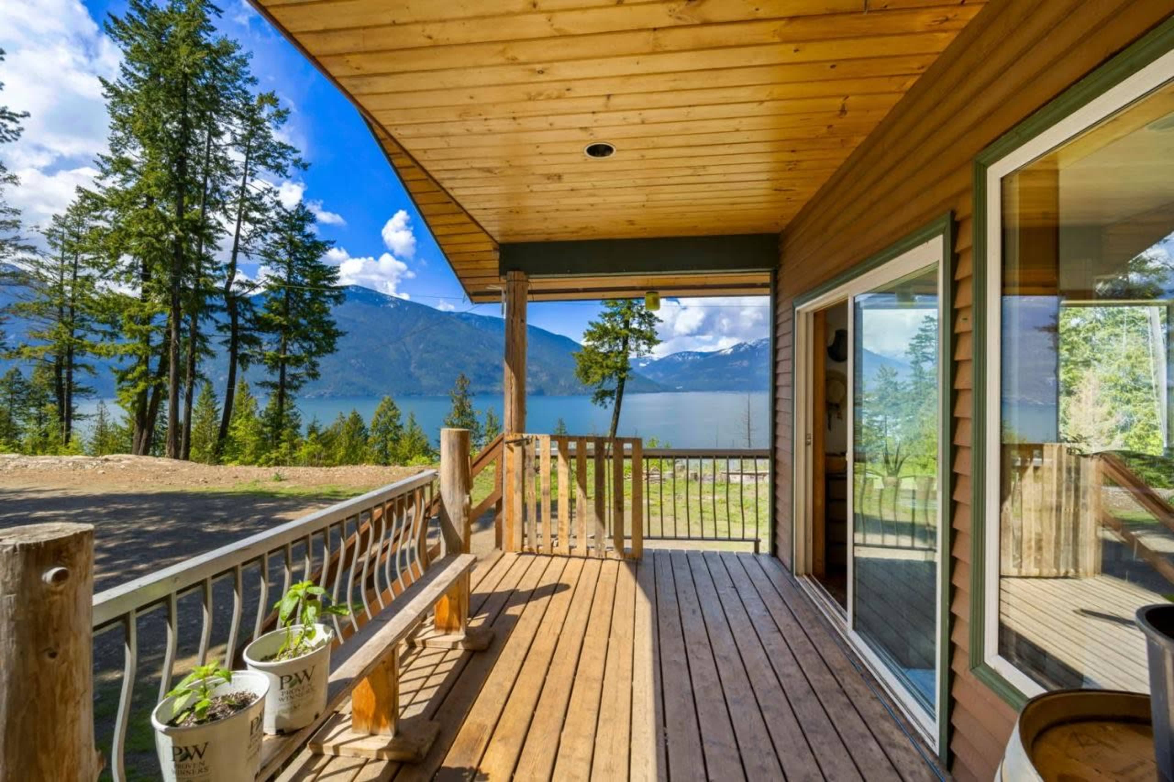 The image shows a wooden deck overlooking a lake, surrounded by tall trees and distant mountains under a partly cloudy sky.