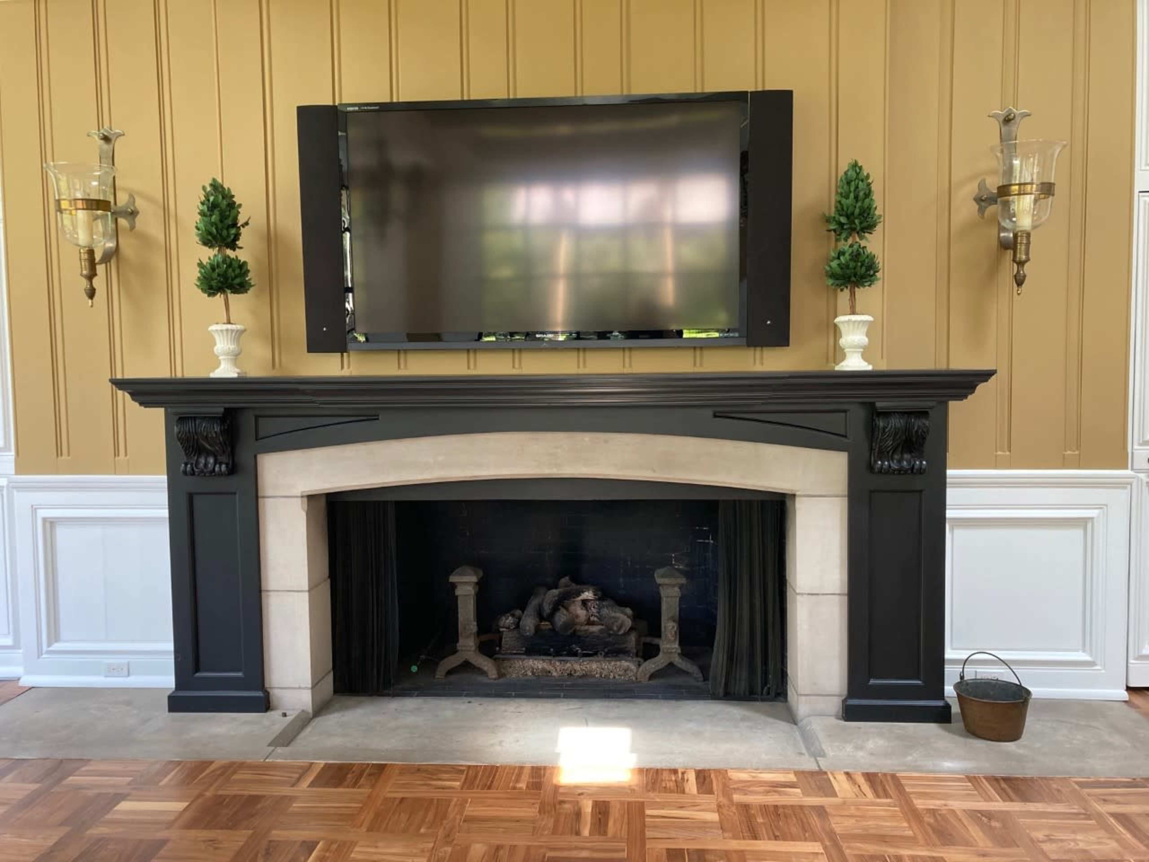 A black mantel with a stone fireplace, flanked by two wall-mounted sconces, and a flat-screen TV mounted above it, set against a gold paneled wall.