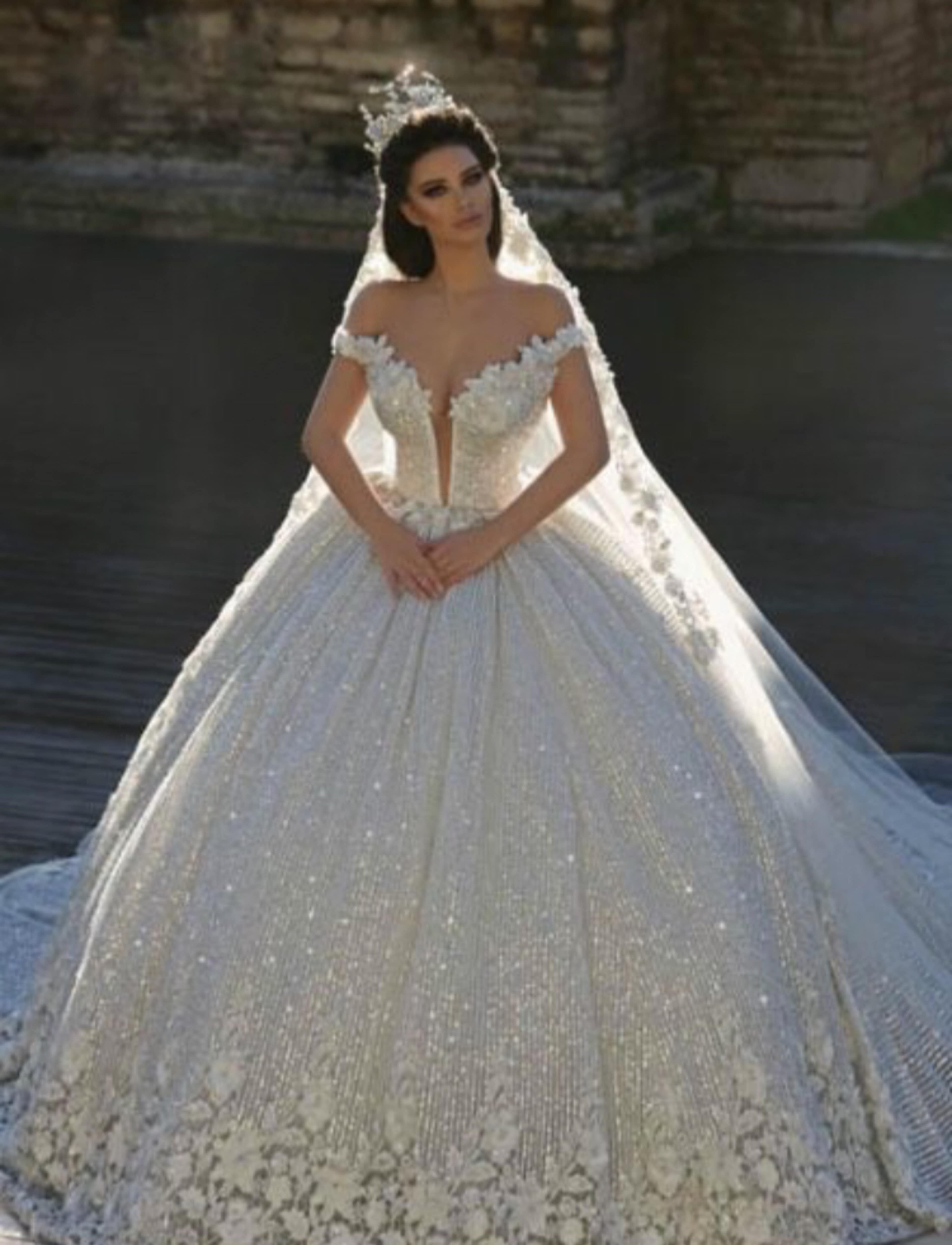 A woman in a lavish off-shoulder wedding gown with intricate floral details and a long veil stands elegantly against a historical backdrop.