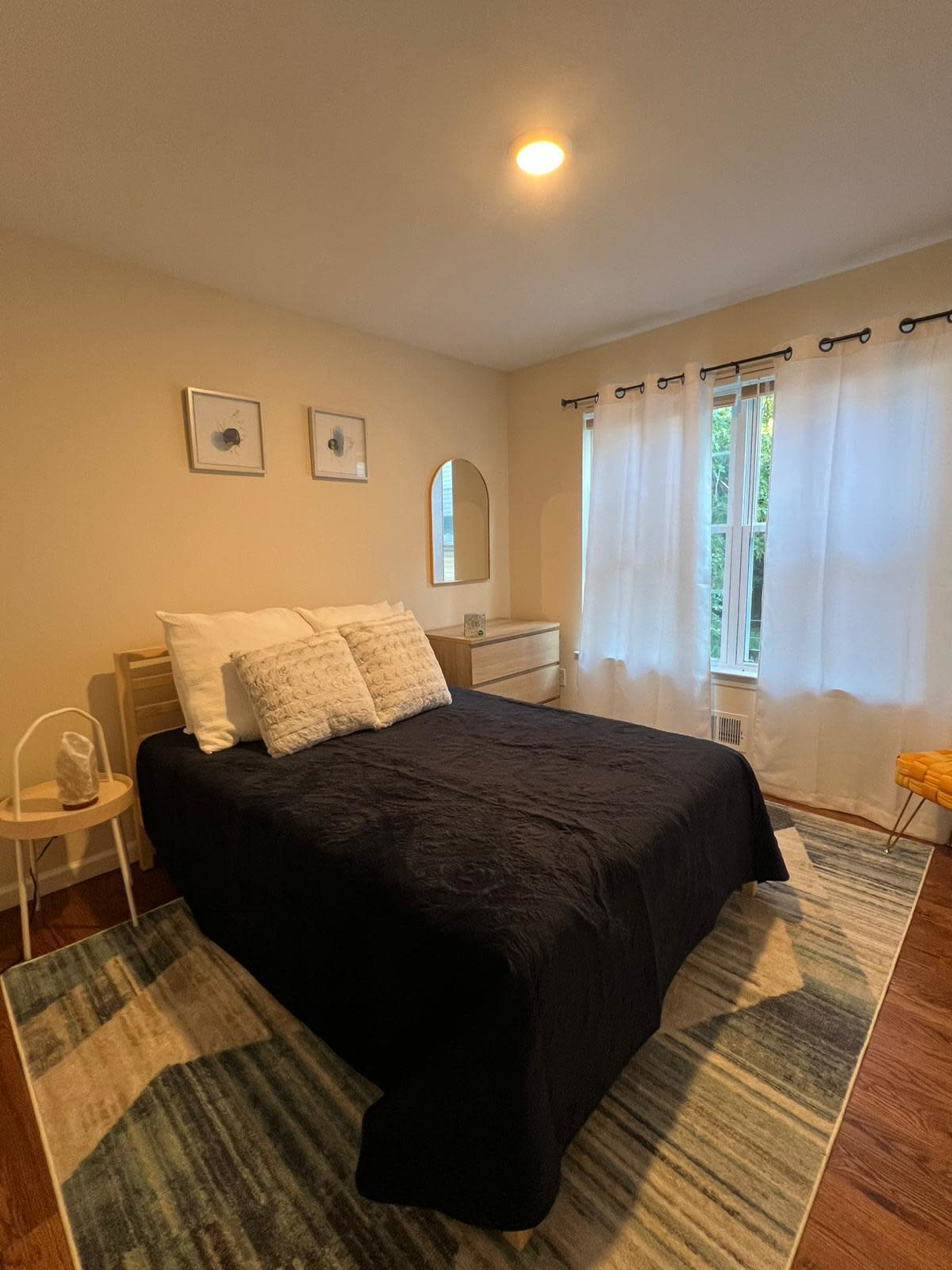 A neatly arranged bedroom with a bed covered in a black quilt, a wooden dresser, and a window dressed with sheer white curtains.