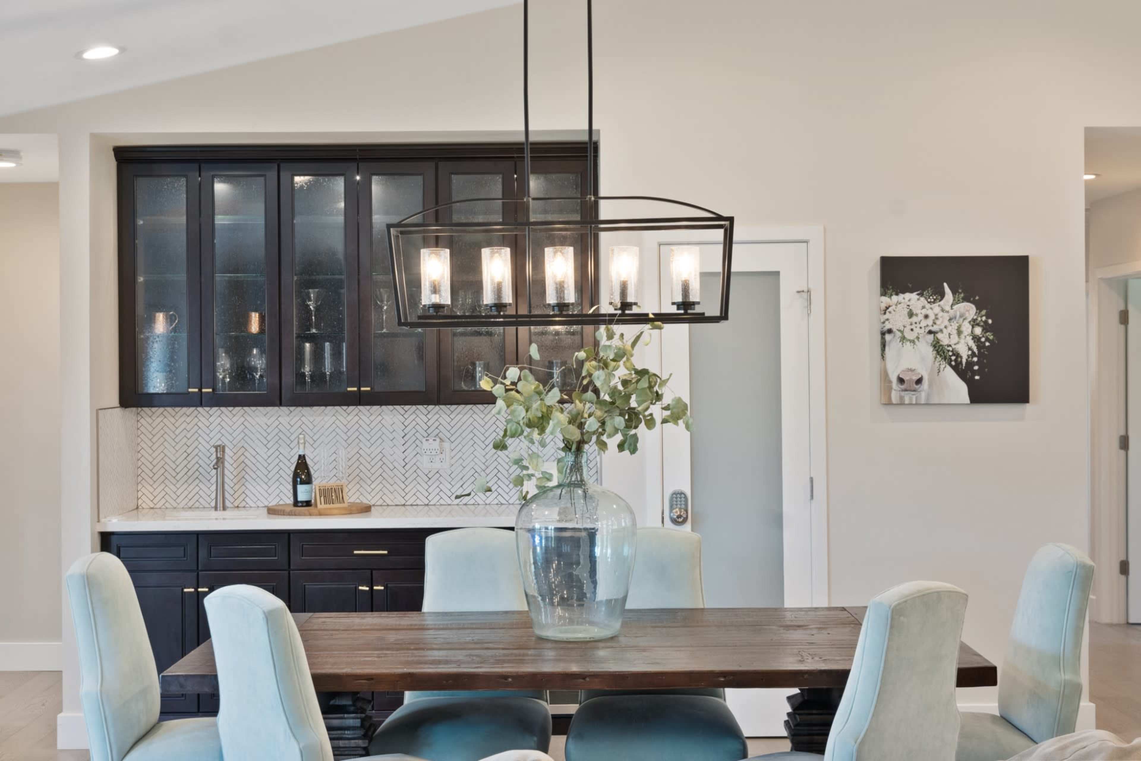A dining area features a wooden table surrounded by upholstered chairs, with a large glass vase of greenery at the center and a modern light fixture above.