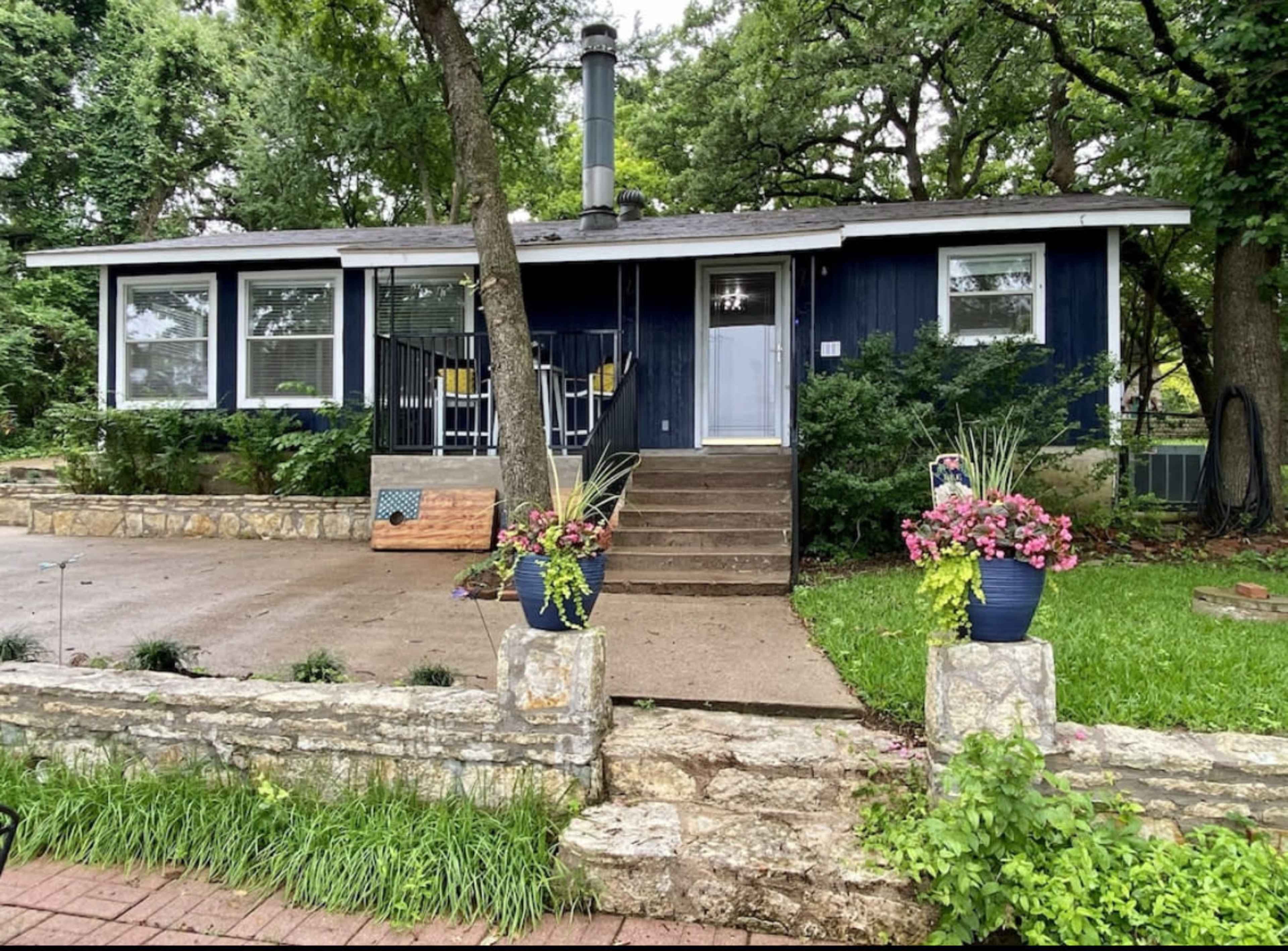 A blue house with a front porch and potted plants is surrounded by trees and a stone wall.