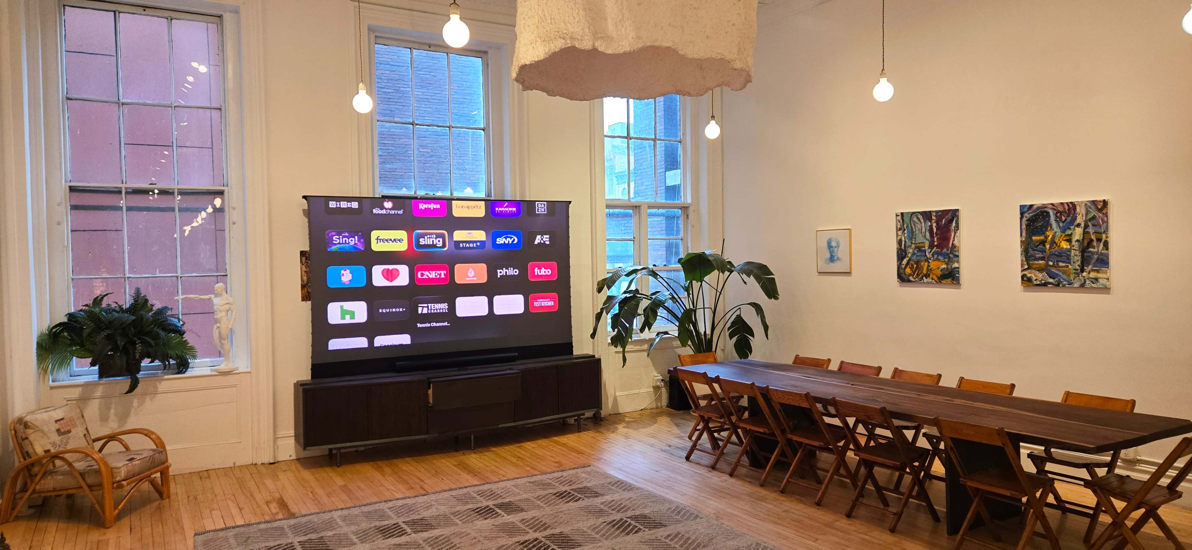 The image shows a bright, spacious room featuring a large TV mounted on the wall, a long wooden dining table with chairs, and several windows allowing natural light to enter, along with a couple of decorative artworks.