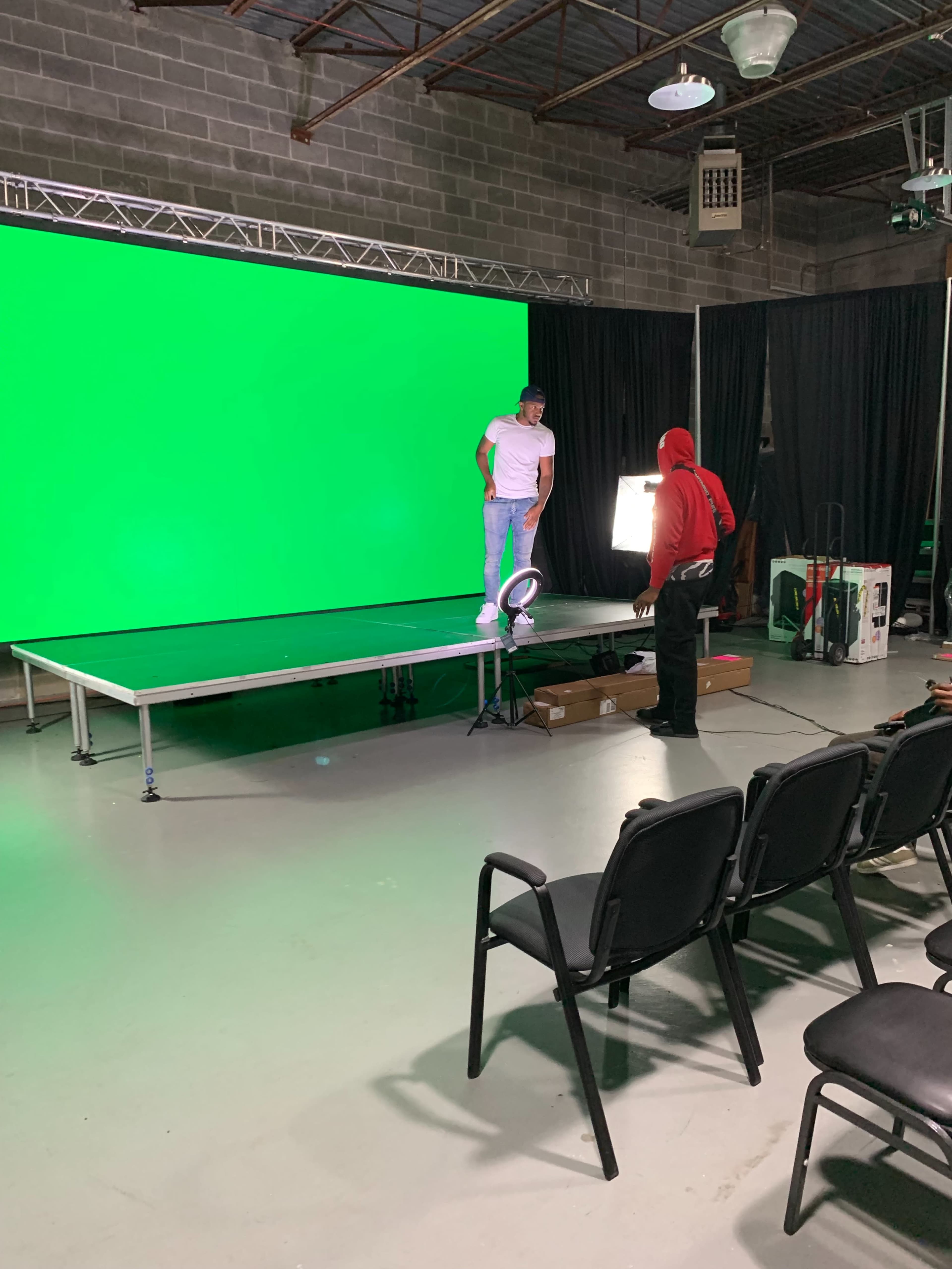 A person in a white shirt stands on a platform in front of a large green screen while another person in a red hoodie adjusts lighting equipment nearby.