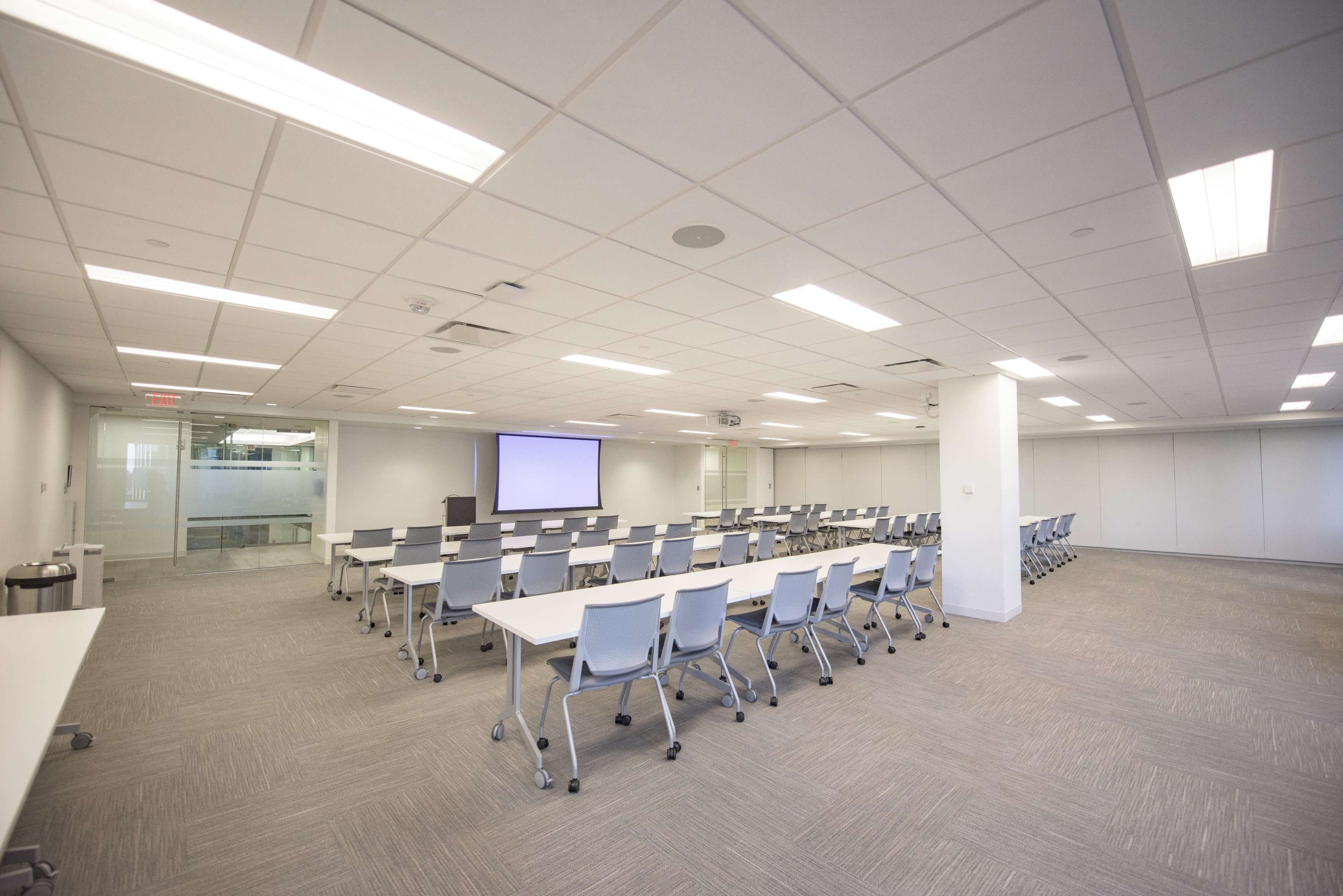A spacious conference room is set up with multiple rows of tables and chairs, accompanied by a screen at the front.