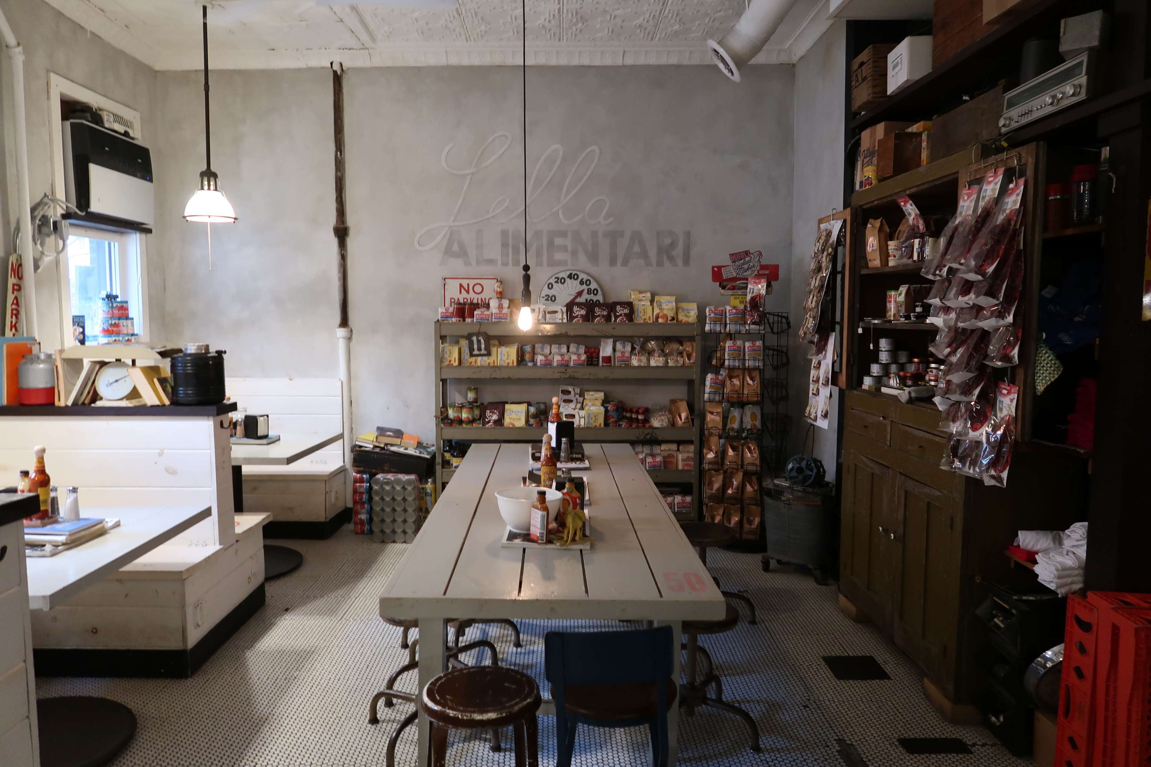 An interior of a rustic café with a communal table, shelving stocked with various food items, and a minimalist design featuring muted colors.