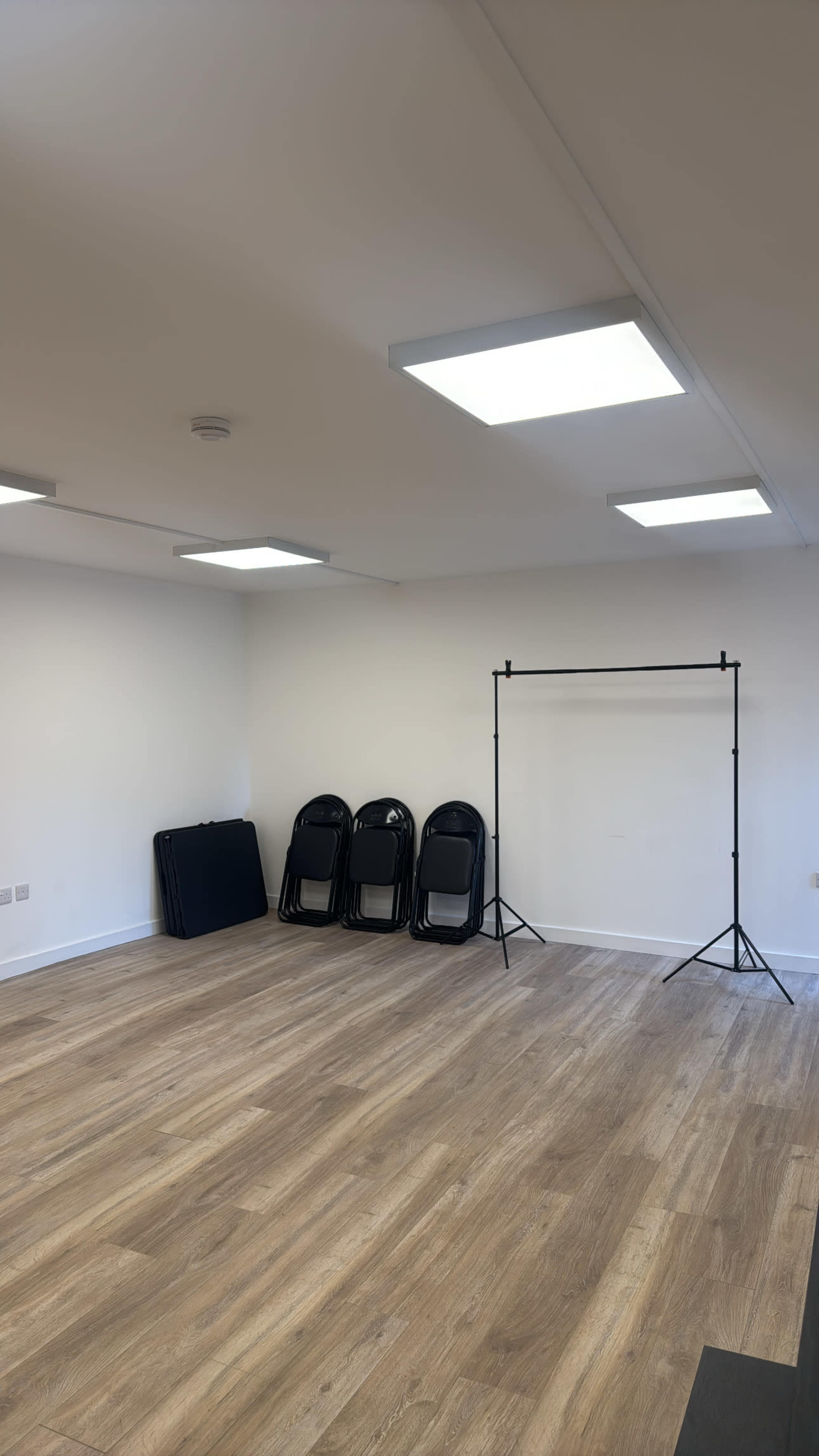 The image shows a vacant room with wooden flooring, five stacked black chairs, and a backdrop stand set up against a white wall.