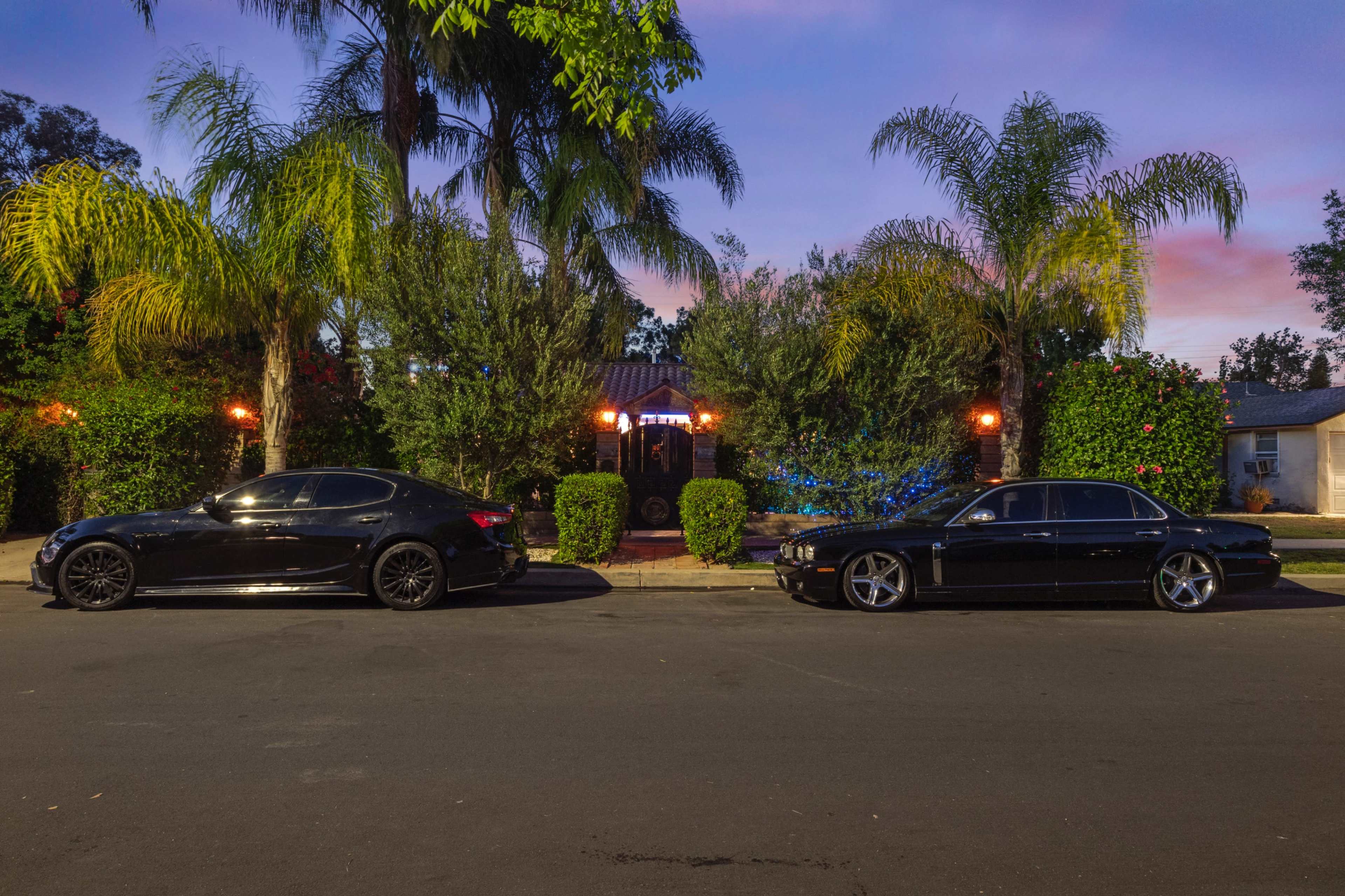 Two black cars are parked on the street in front of a house surrounded by palm trees and well-maintained greenery at dusk.