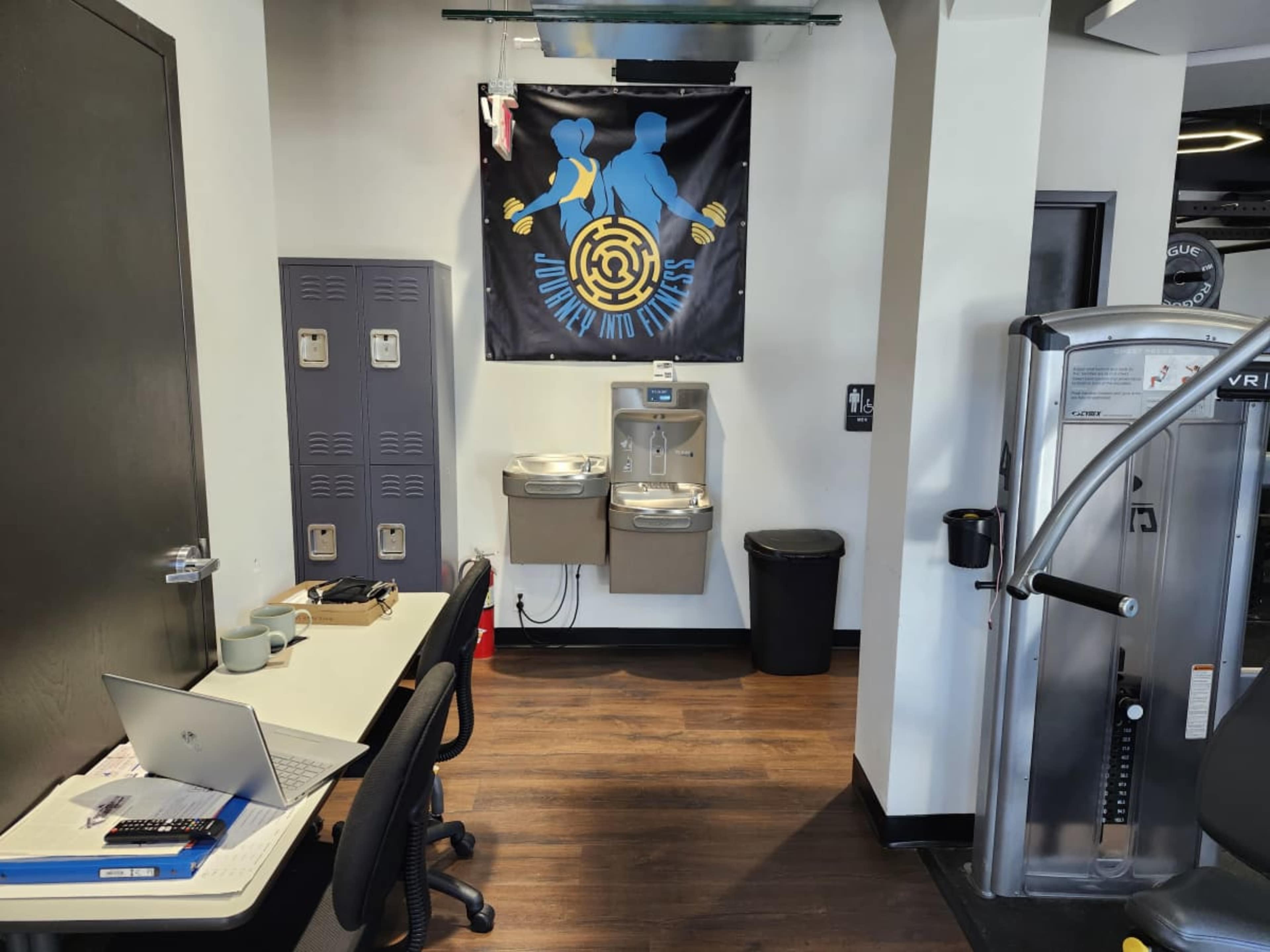 The image shows a gym area featuring a water fountain, lockers, a table with a laptop, and fitness equipment in a well-lit space.