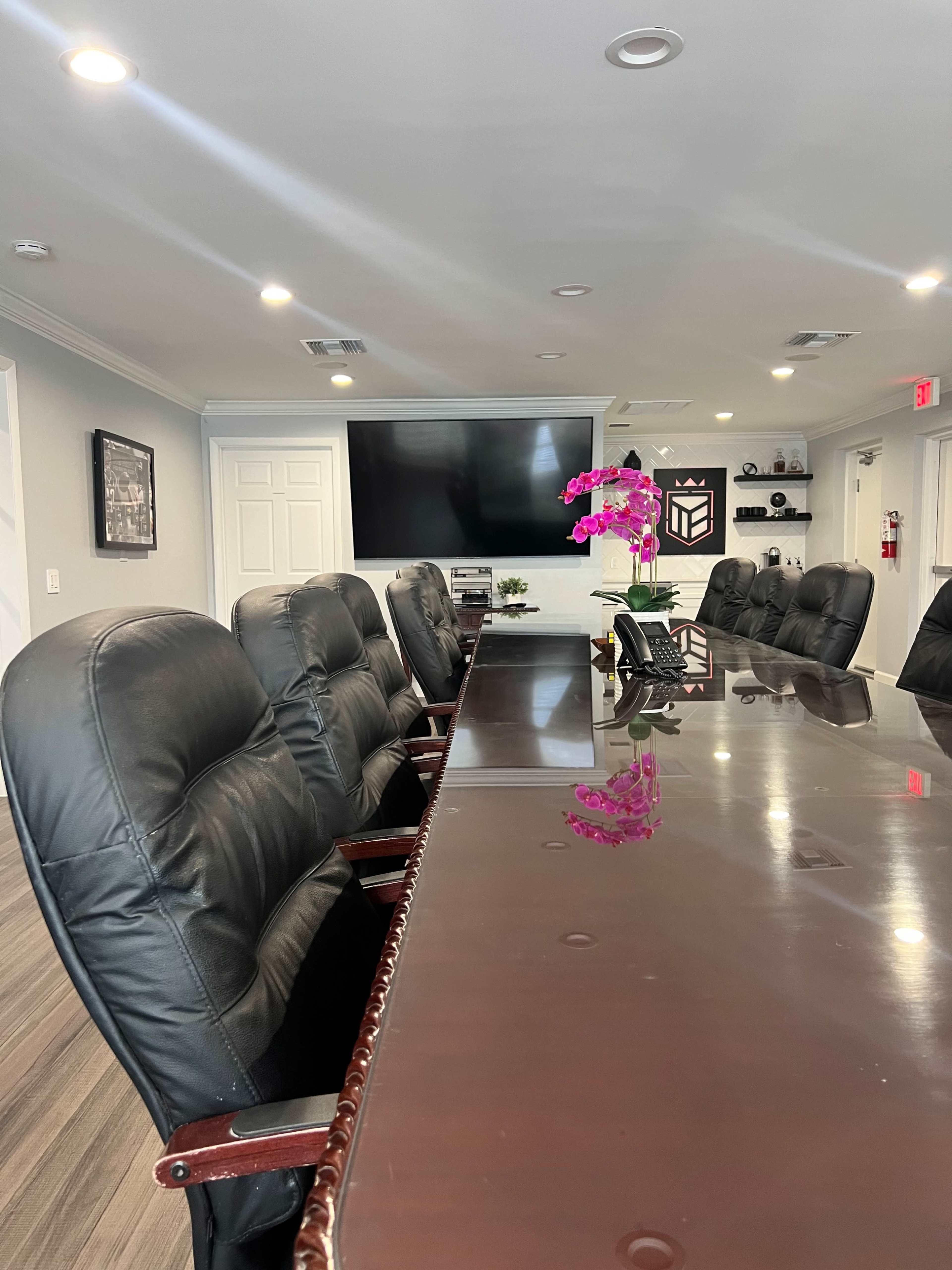 A conference room equipped with a long table, black office chairs, and a large wall-mounted screen at one end.