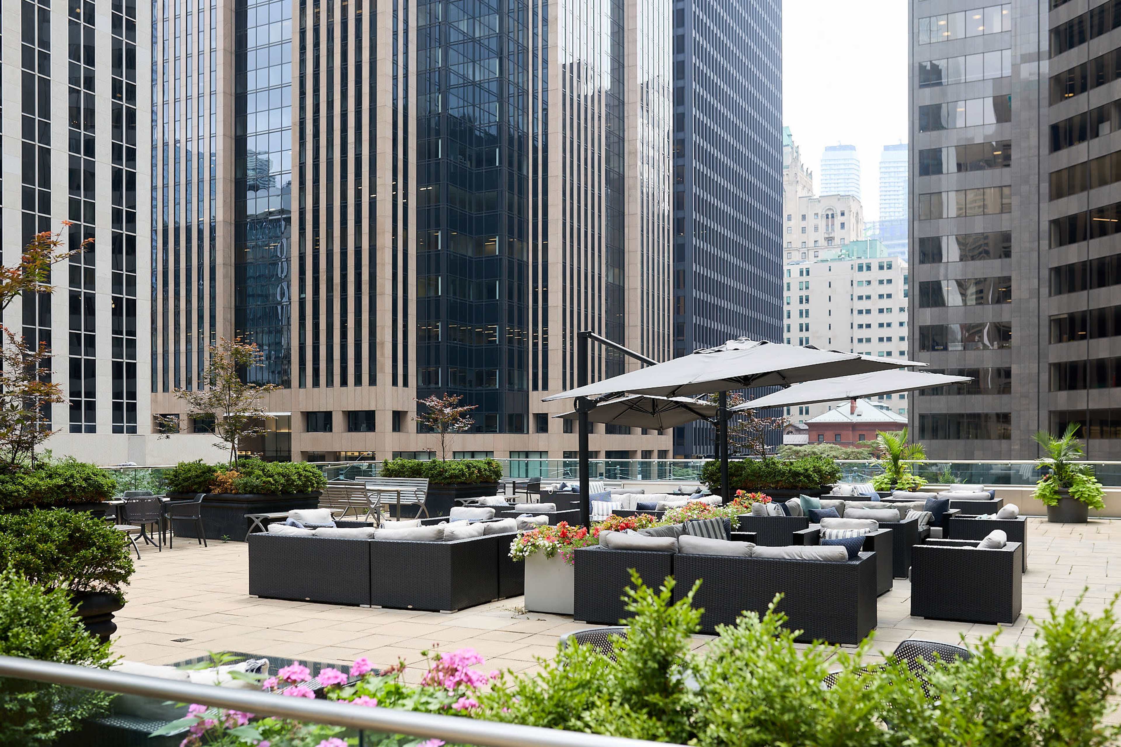 The image shows a modern rooftop terrace with seating arrangements, plants, and an umbrella, surrounded by tall glass skyscrapers.
