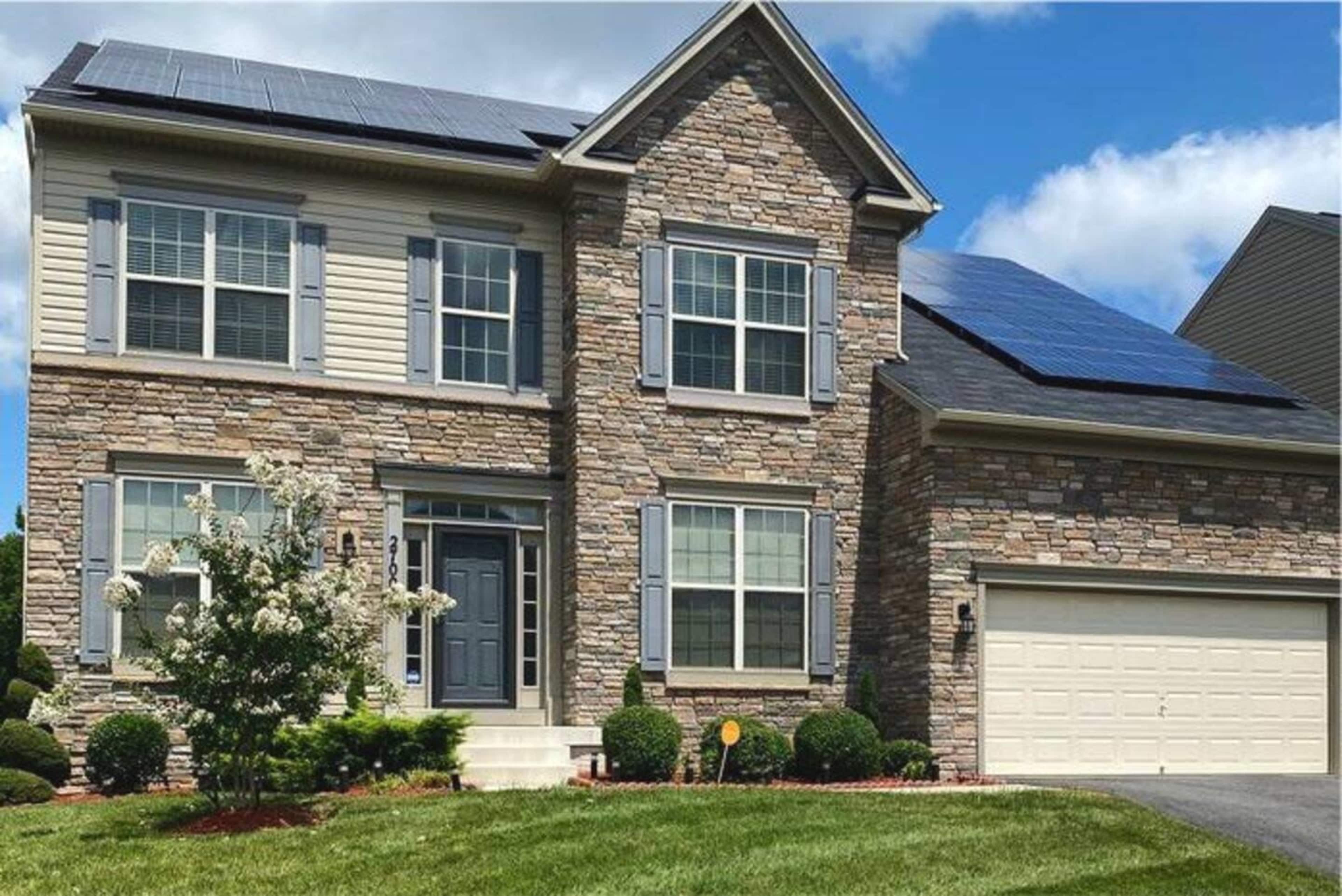 The image shows a two-story stone-clad house with solar panels on the roof and a two-car garage.