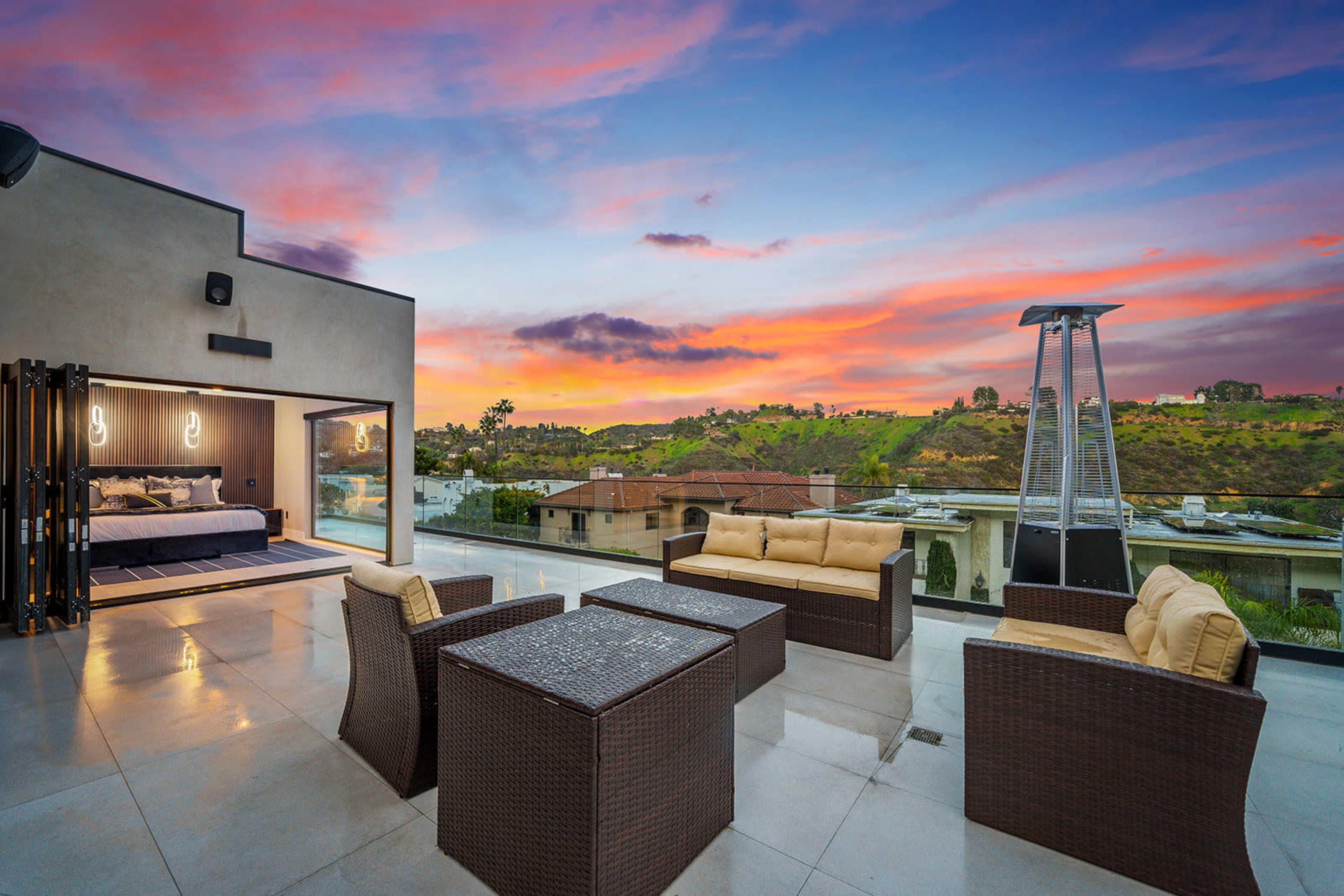 A modern rooftop terrace features outdoor seating and a view of the sunset over hills and houses.