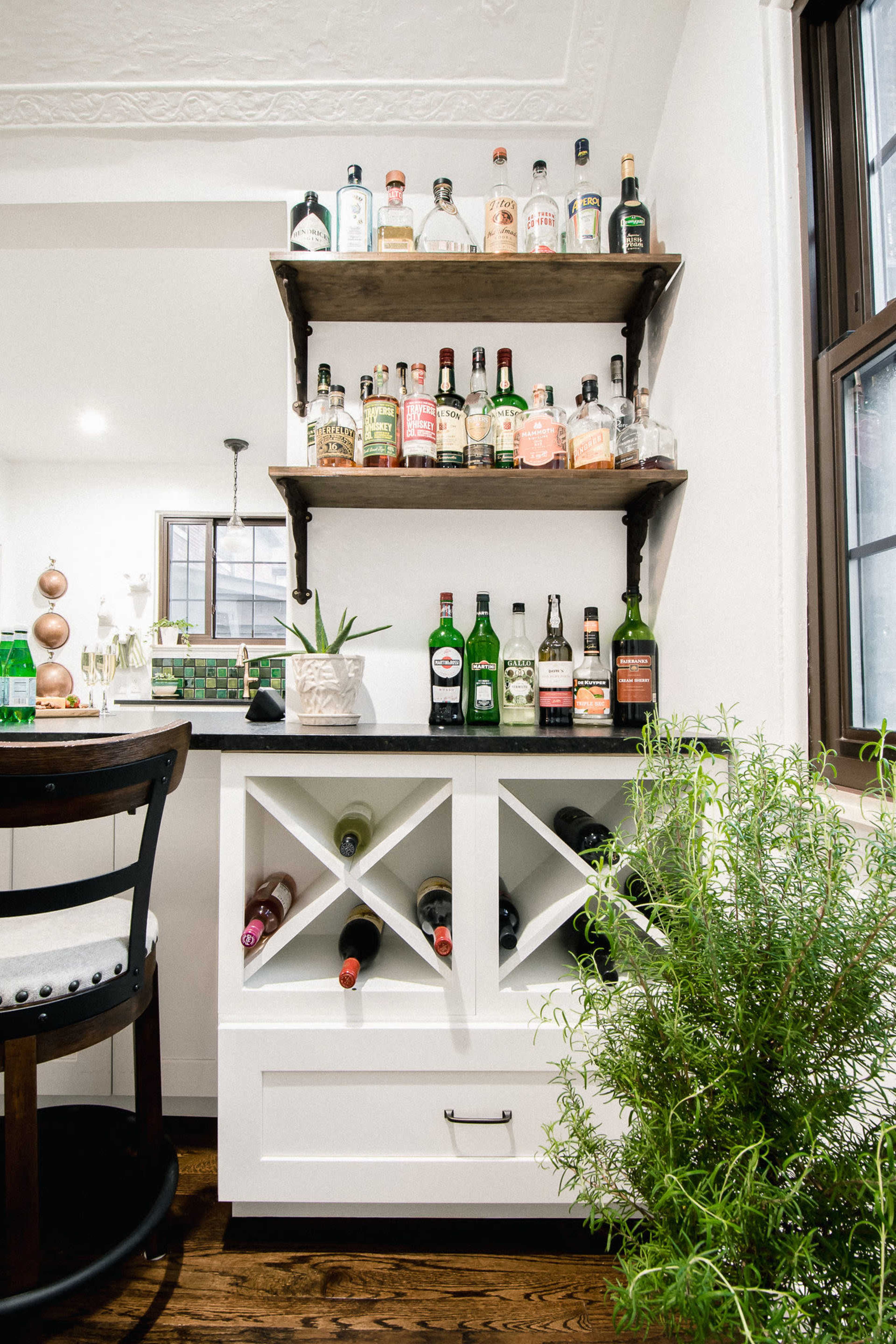A home bar area featuring a white cabinet with diagonal wine bottle storage, a drawer, a green plant nearby, and shelves above displaying various liquor bottles.