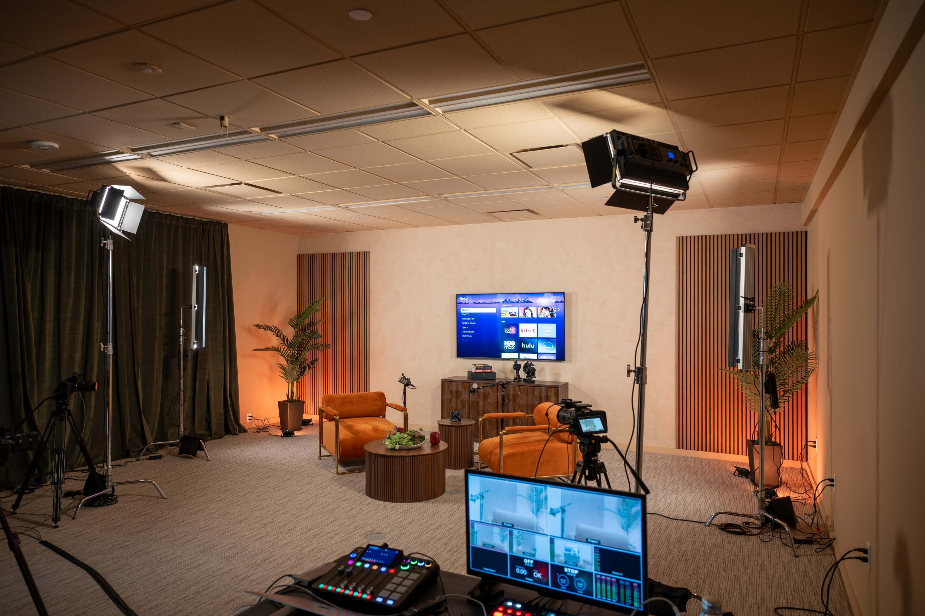 A modern video production studio features two orange chairs, a large screen displaying graphics, and various lighting and camera equipment arranged around the room.