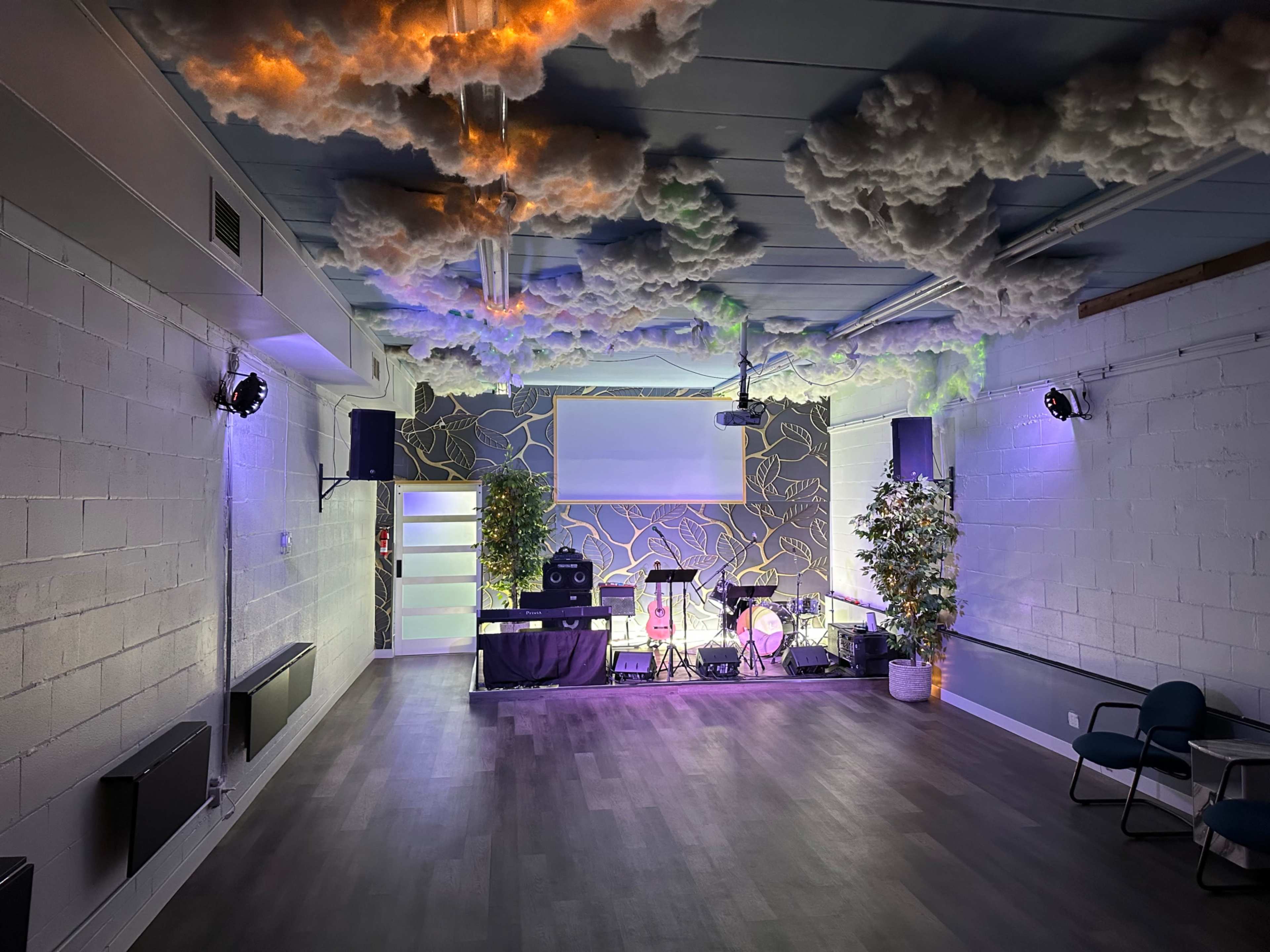 The image shows an indoor event space with a stage set up, featuring musical instruments, decorative plants, and a ceiling designed to resemble clouds.