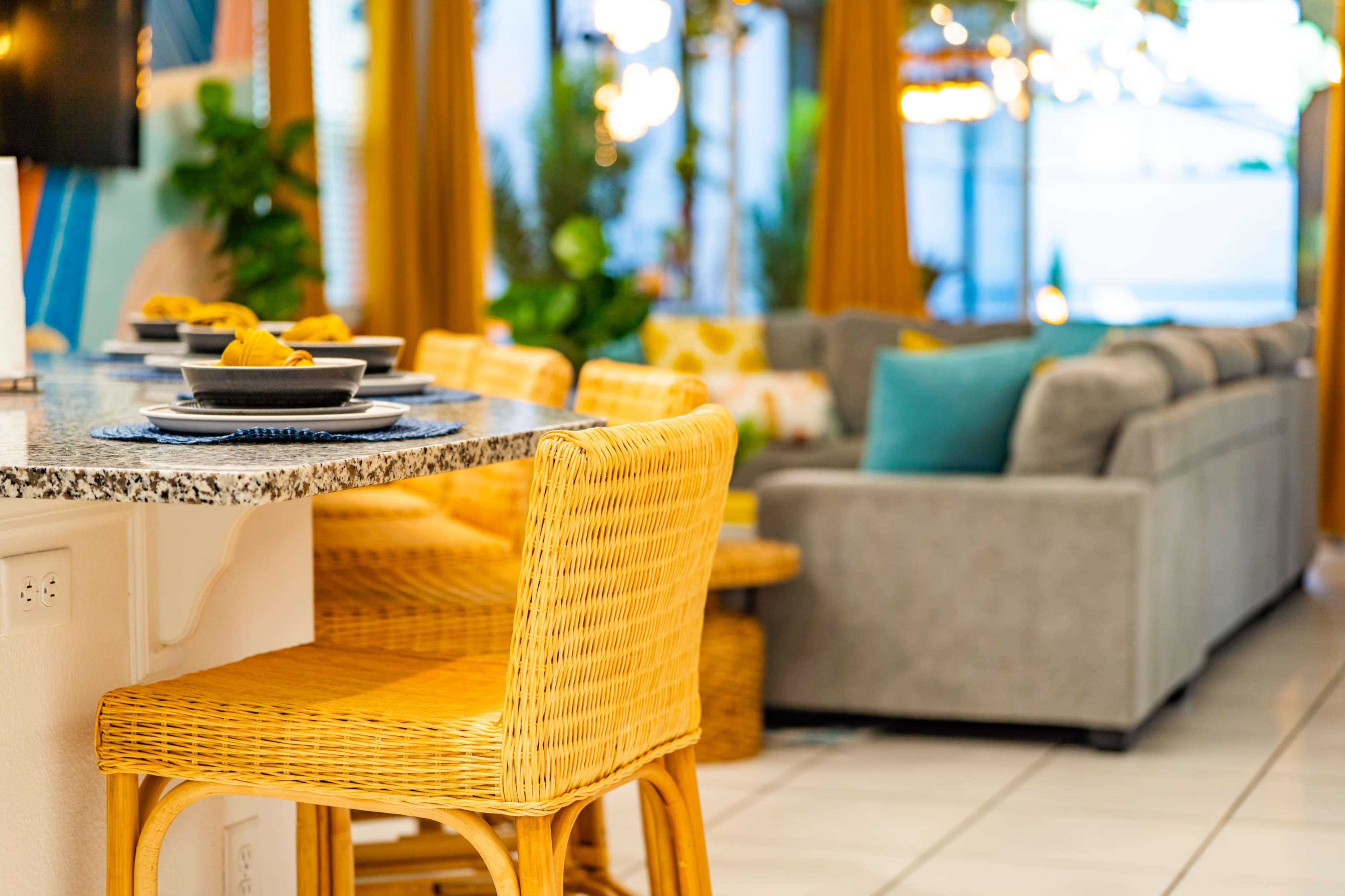 A modern dining area with a granite countertop, wicker bar stools, and a gray sofa in the background.
