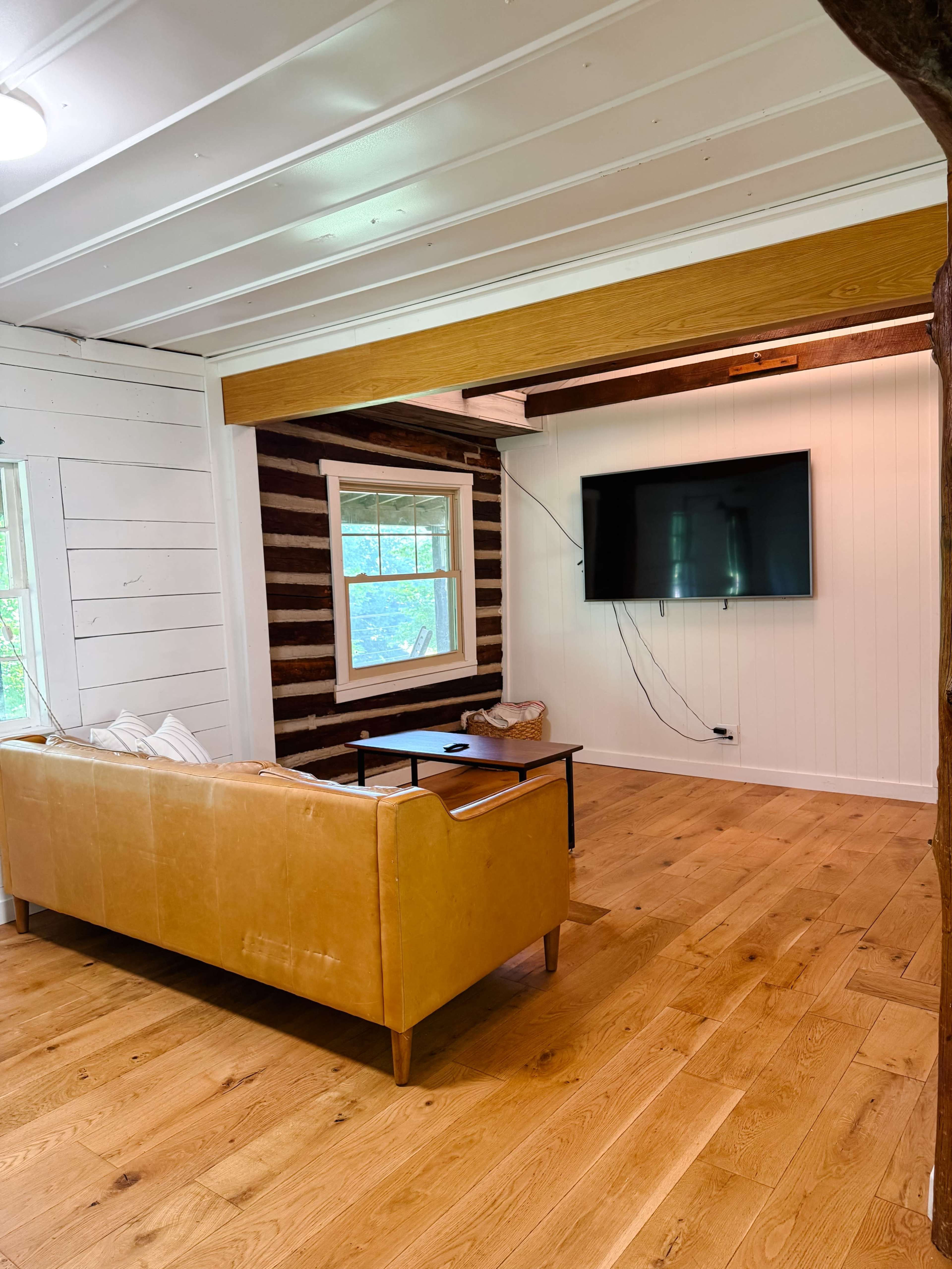 The image shows a living room with a tan leather sofa, a black table, and a mounted television on a white wood-paneled wall.