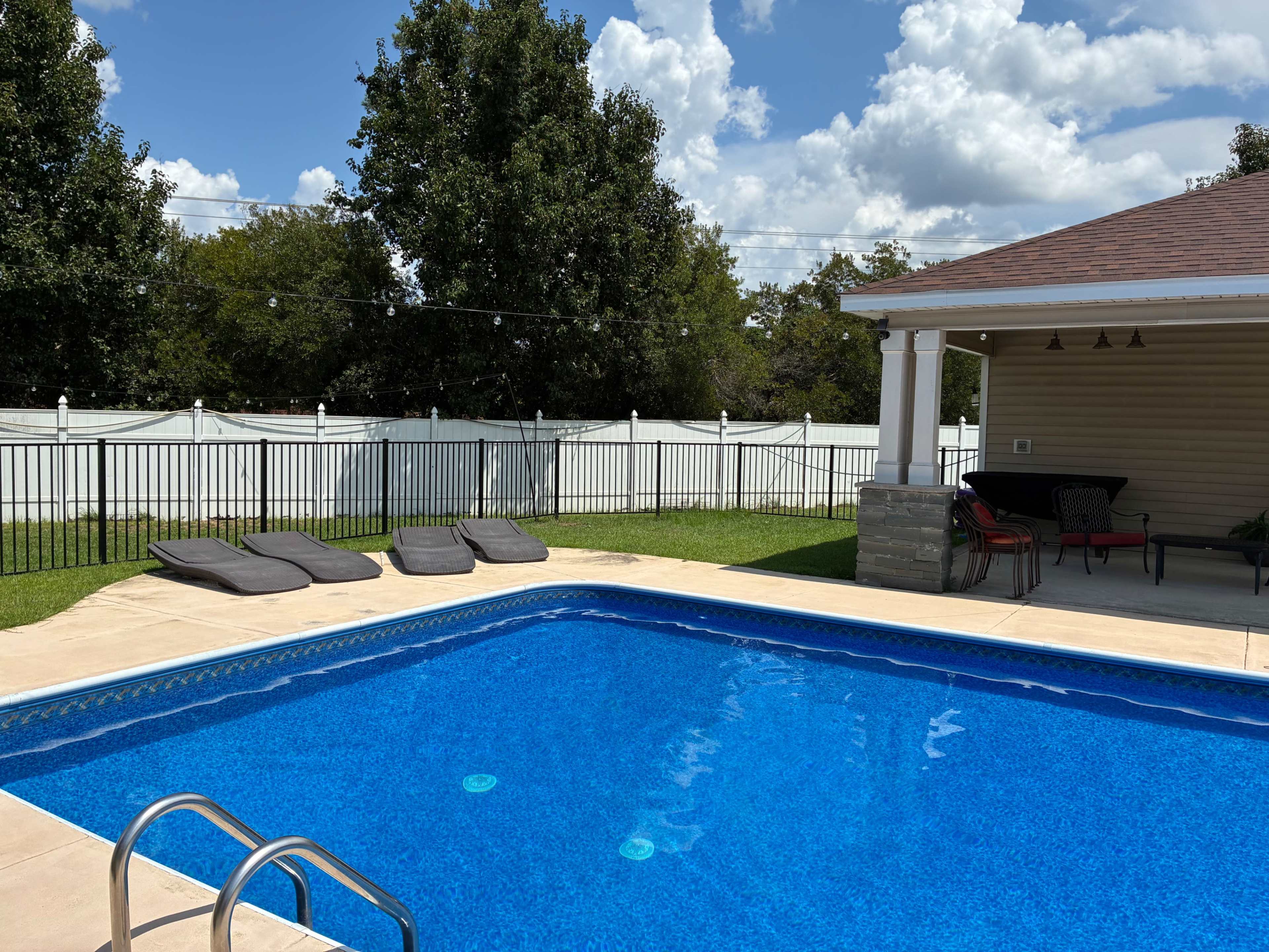 The image shows a swimming pool with three lounge chairs nearby, surrounded by a green lawn and a white fence under a partly cloudy sky.