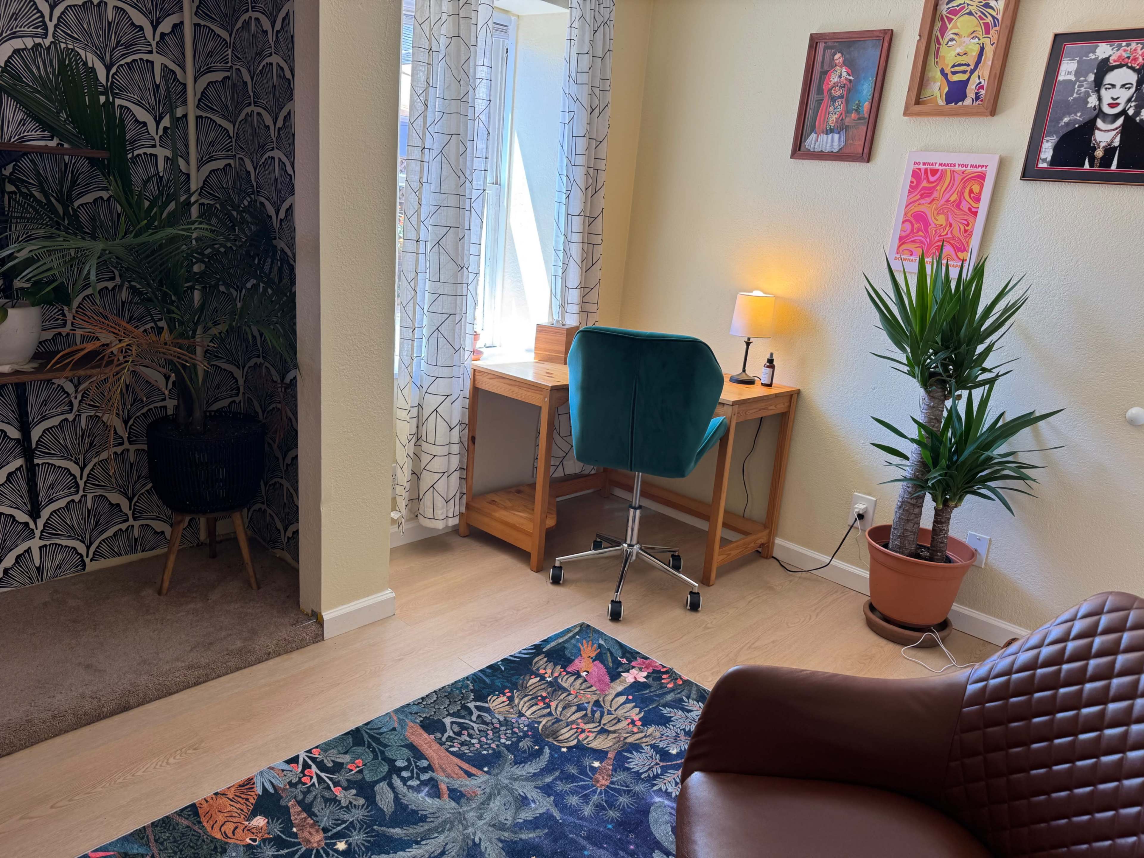 The image shows a room with a small desk and chair, colorful artwork on the walls, a potted plant, and a patterned rug.