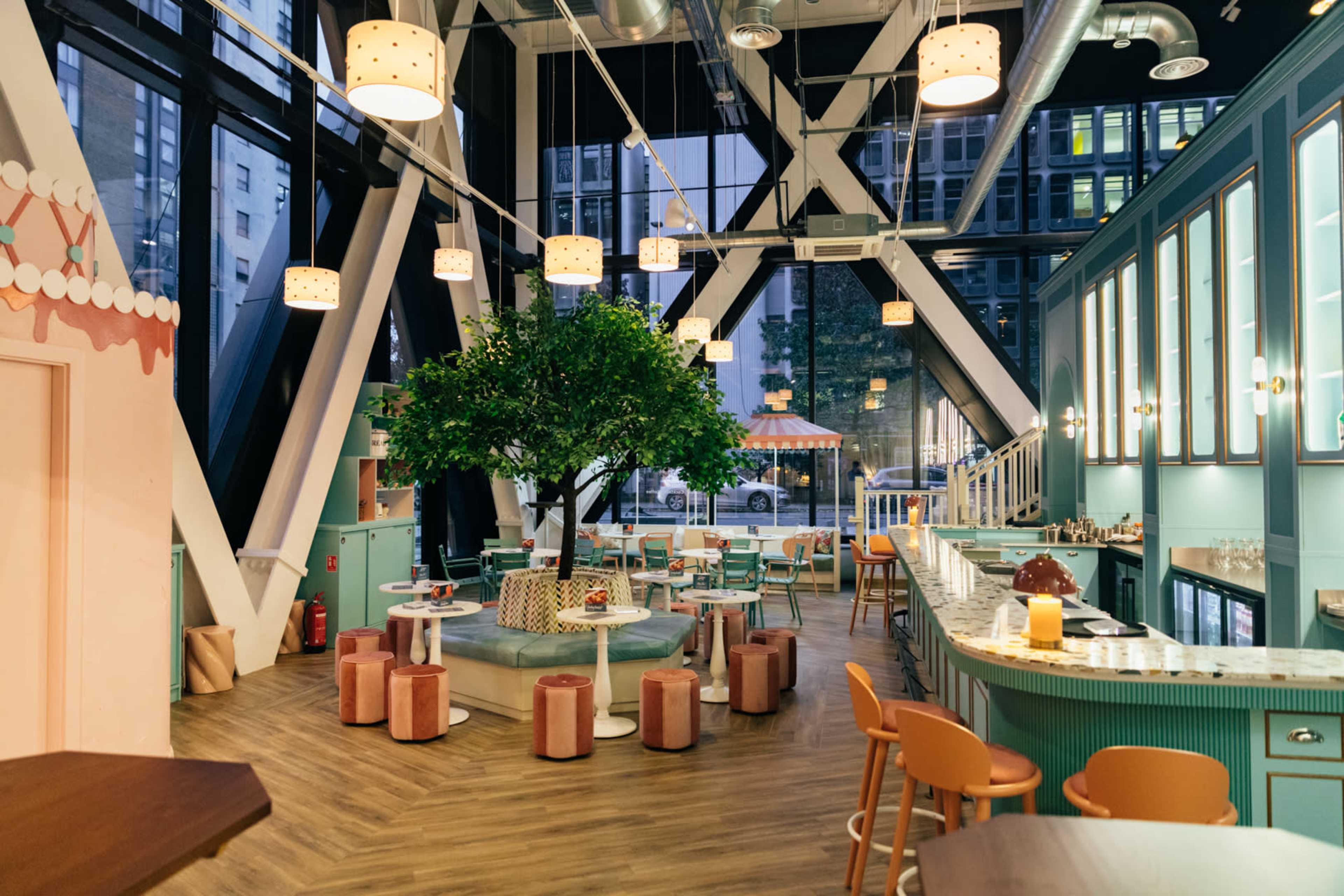The image shows a modern cafe with large windows, featuring a prominent tree in the center, surrounded by colorful seating and a bar area.