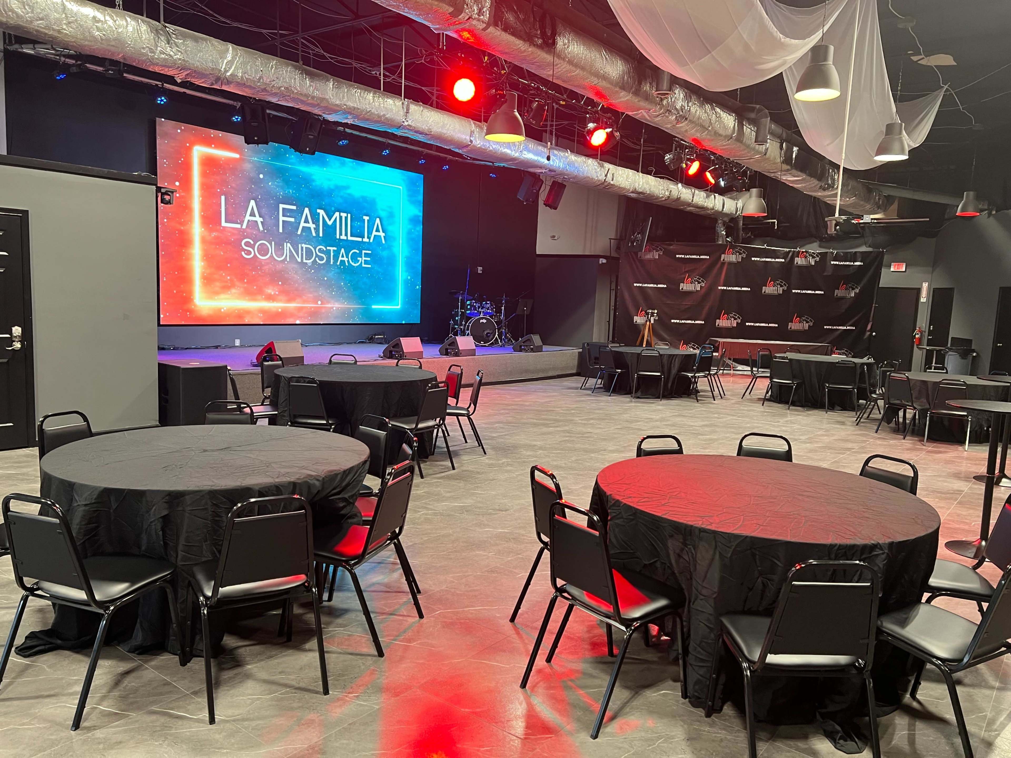 A spacious event venue featuring round black tables and chairs, with a stage in the background displaying a colorful sign that reads "LA FAMILIA SOUNDSTAGE."