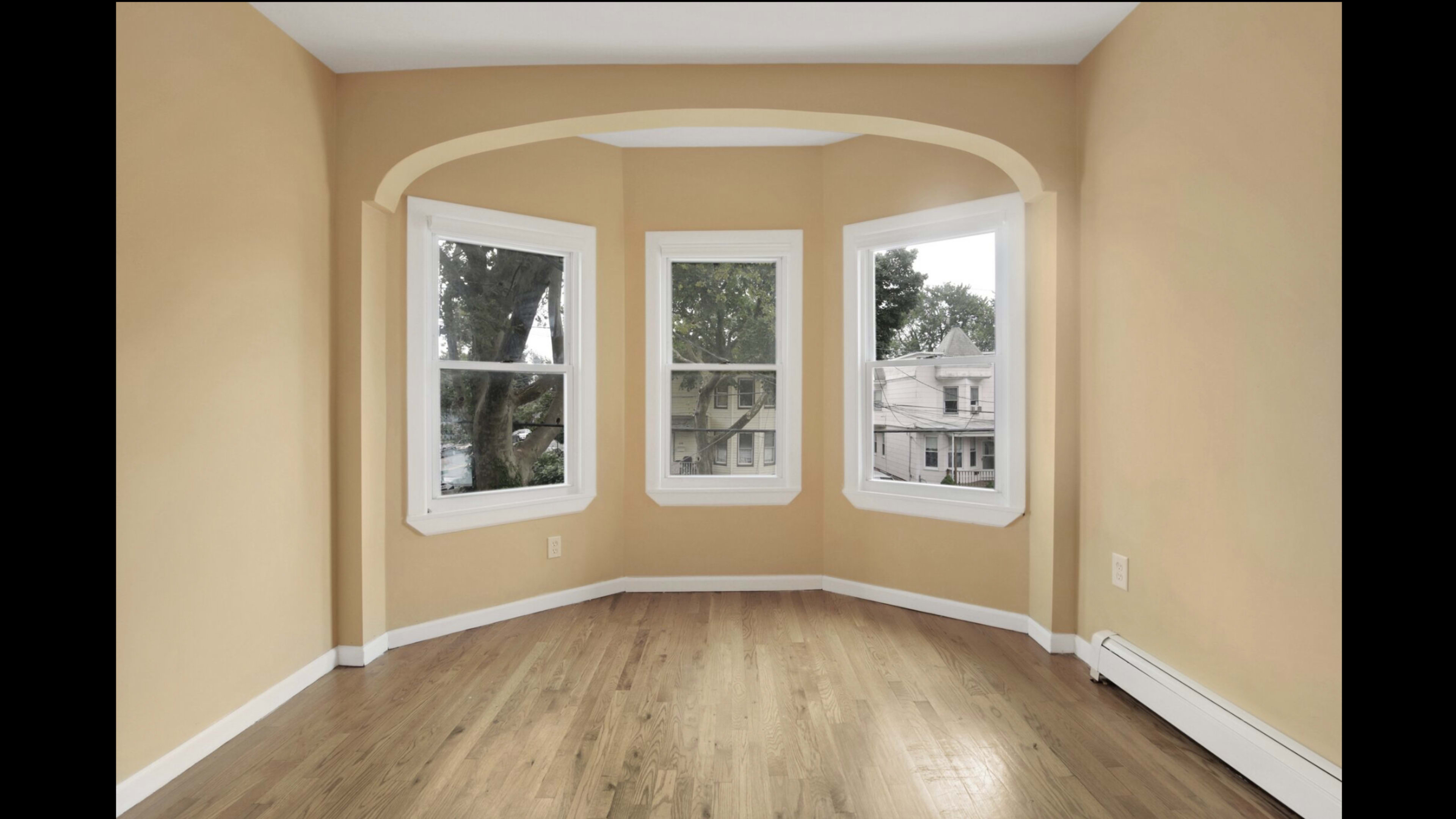The image shows an empty room with light orange walls and three large windows that provide natural light.