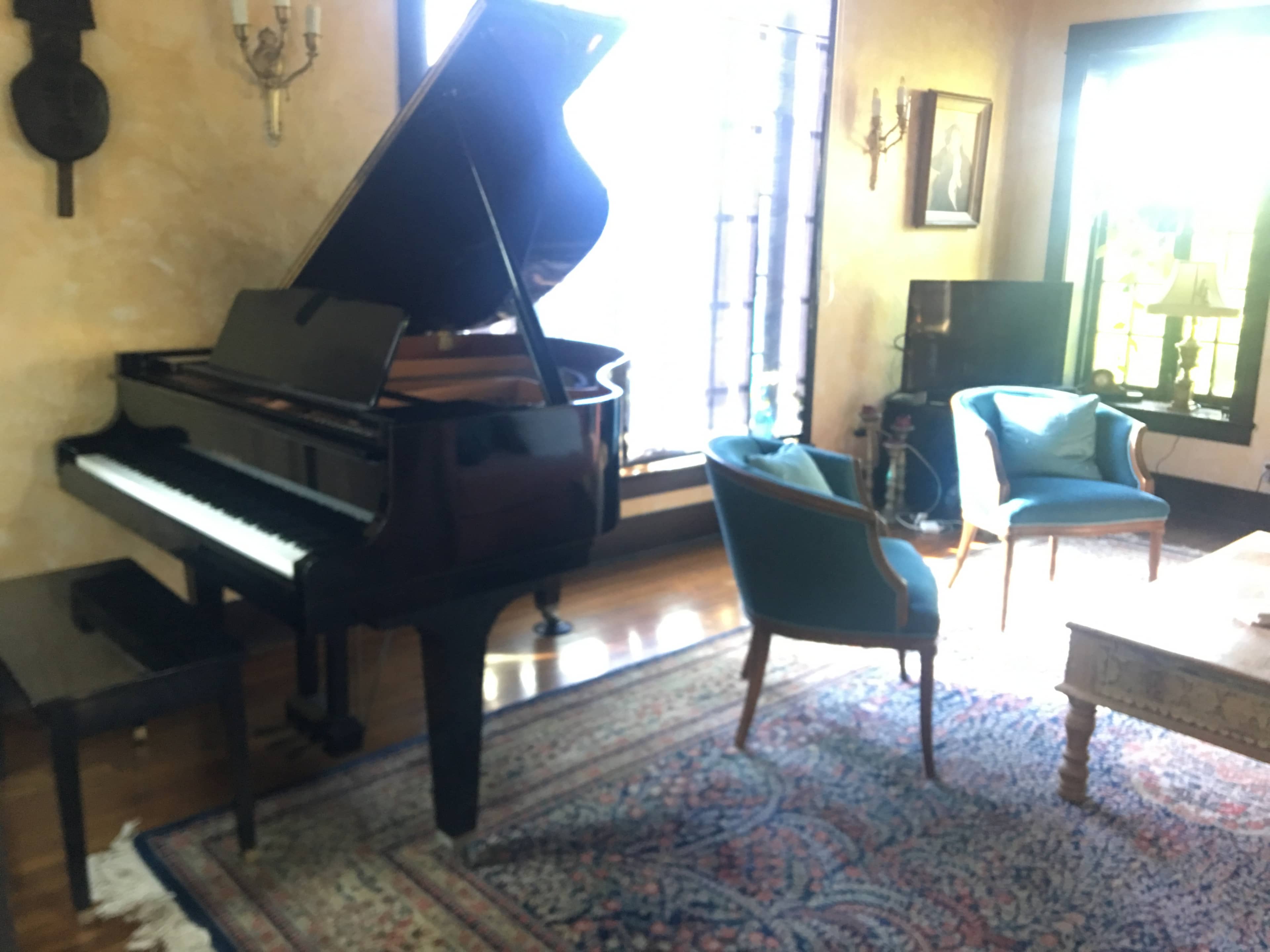 A grand piano stands next to a pair of upholstered chairs in a well-lit living room with large windows and a patterned rug.