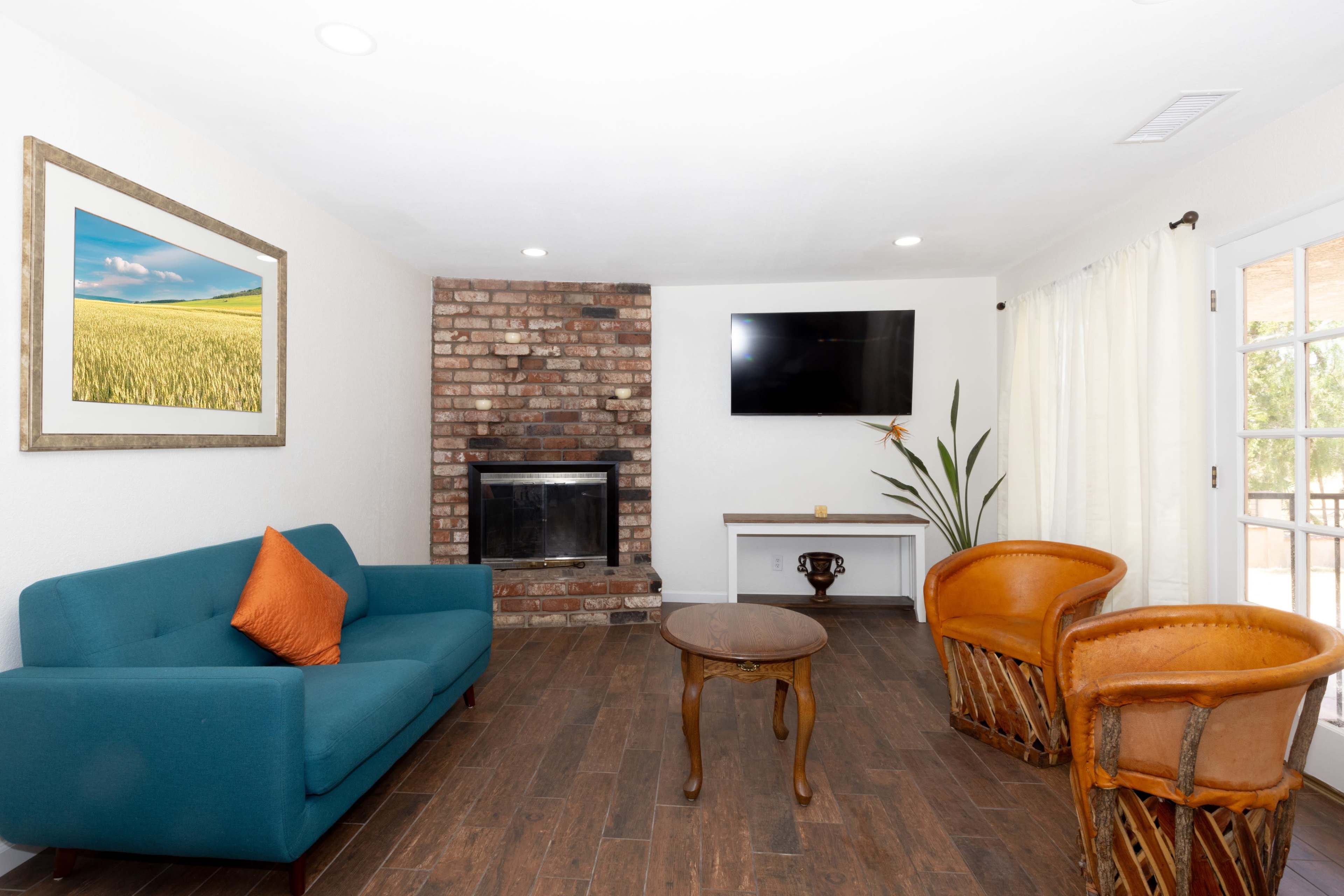 A living room with a turquoise couch, two wooden chairs, a brick fireplace, and a television mounted on the wall.