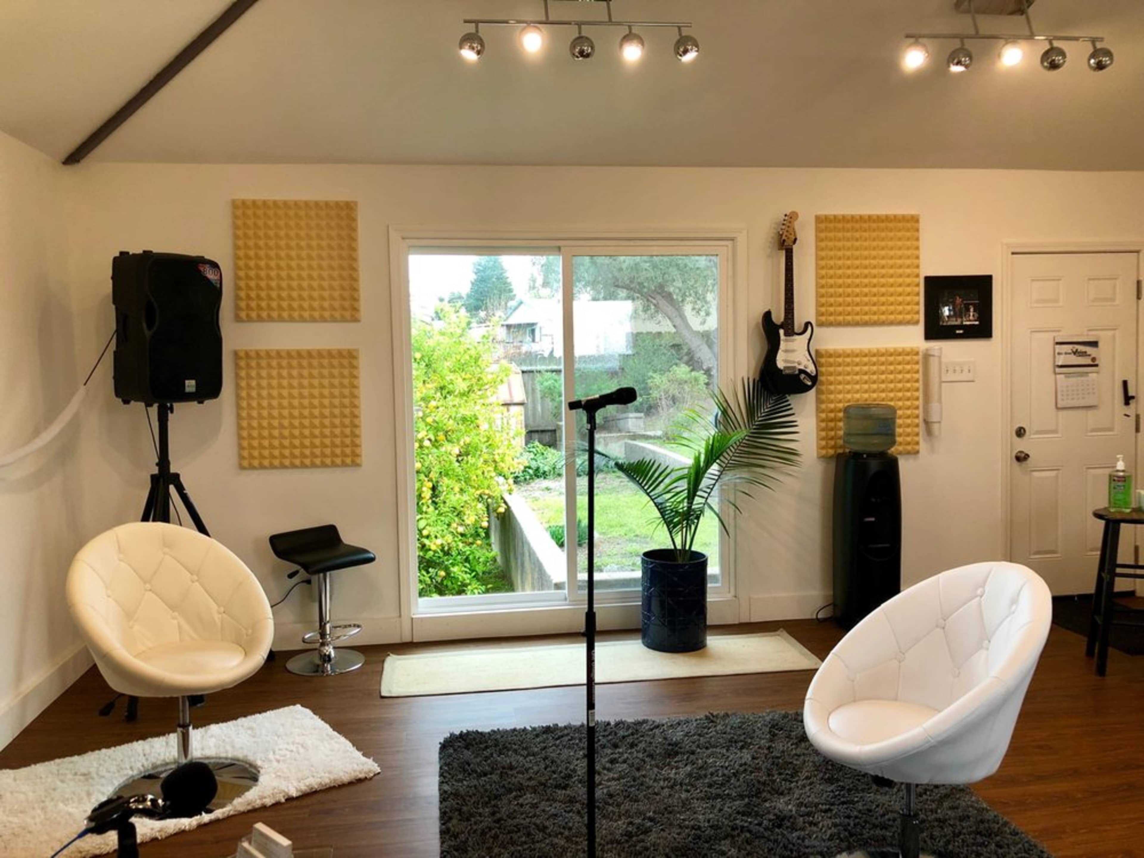 The room features two white chairs, a microphone stand, and a large window overlooking greenery, with acoustic panels on the walls.