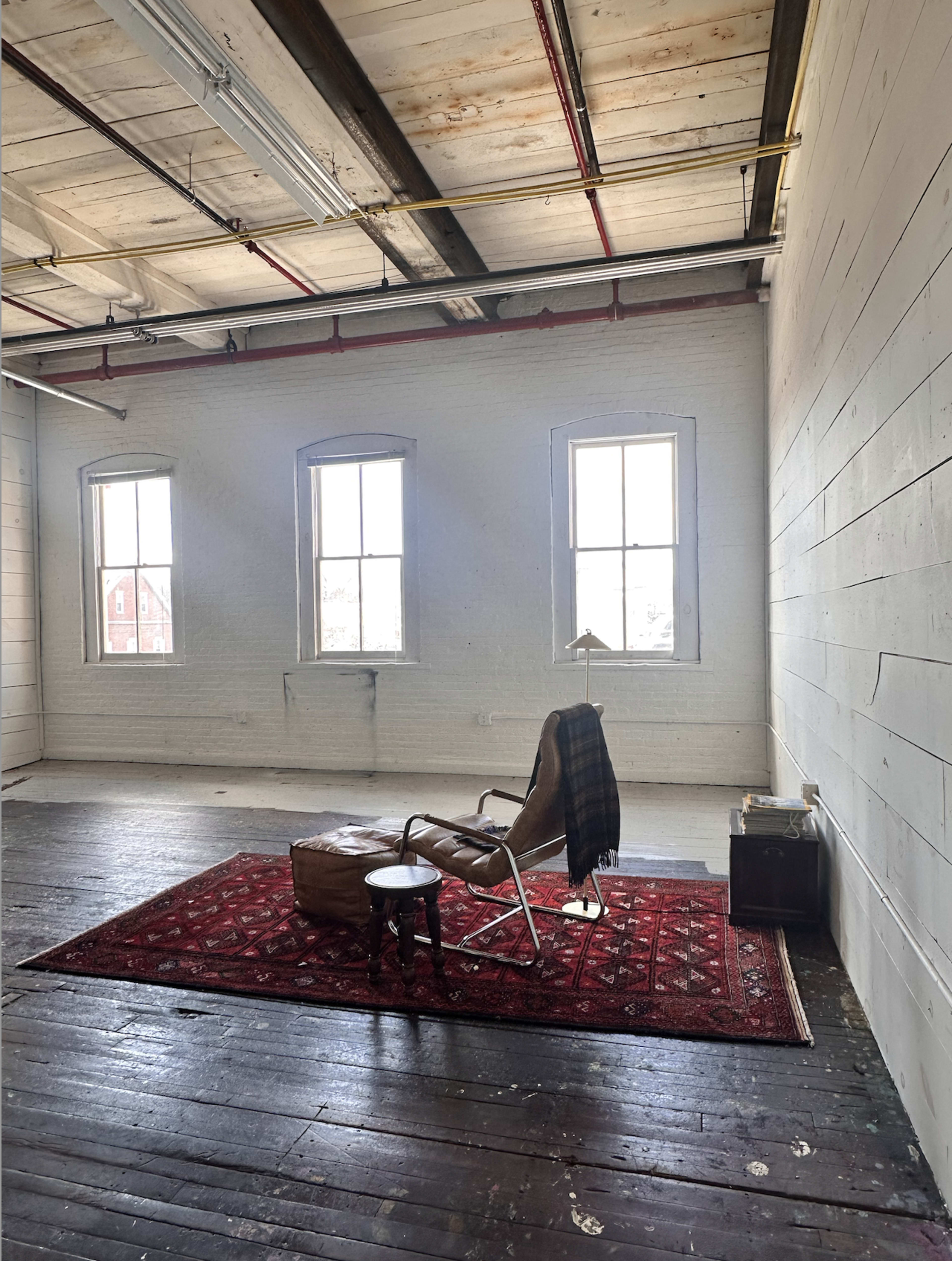 The image shows a spacious room with exposed wooden beams, three large windows allowing natural light, a vintage armchair, a small round table, a rug, and a leather ottoman.