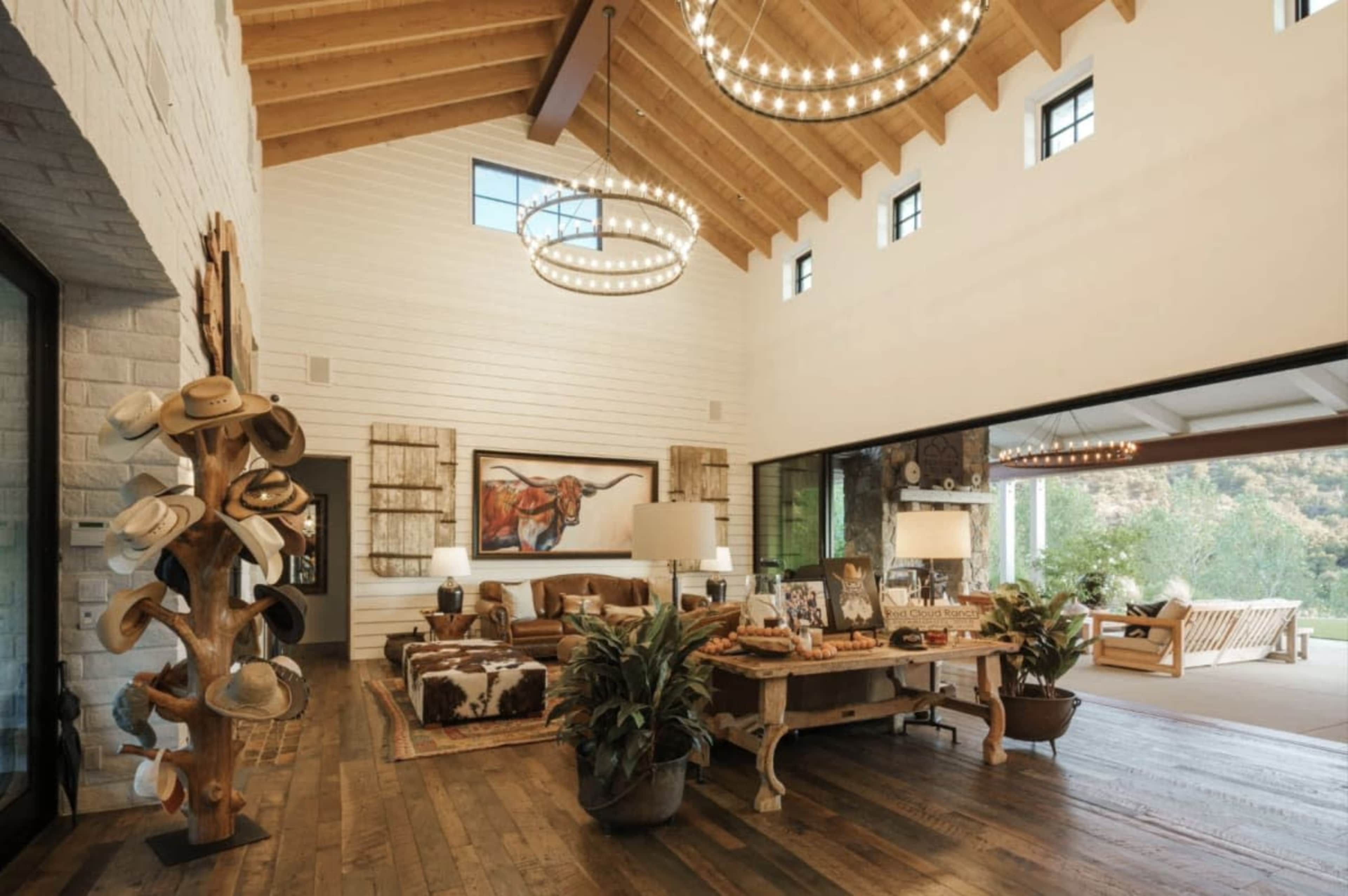 The image shows a spacious living room with high ceilings, featuring a large artwork, a wooden table with decor, and a hat display on a wooden stand.
