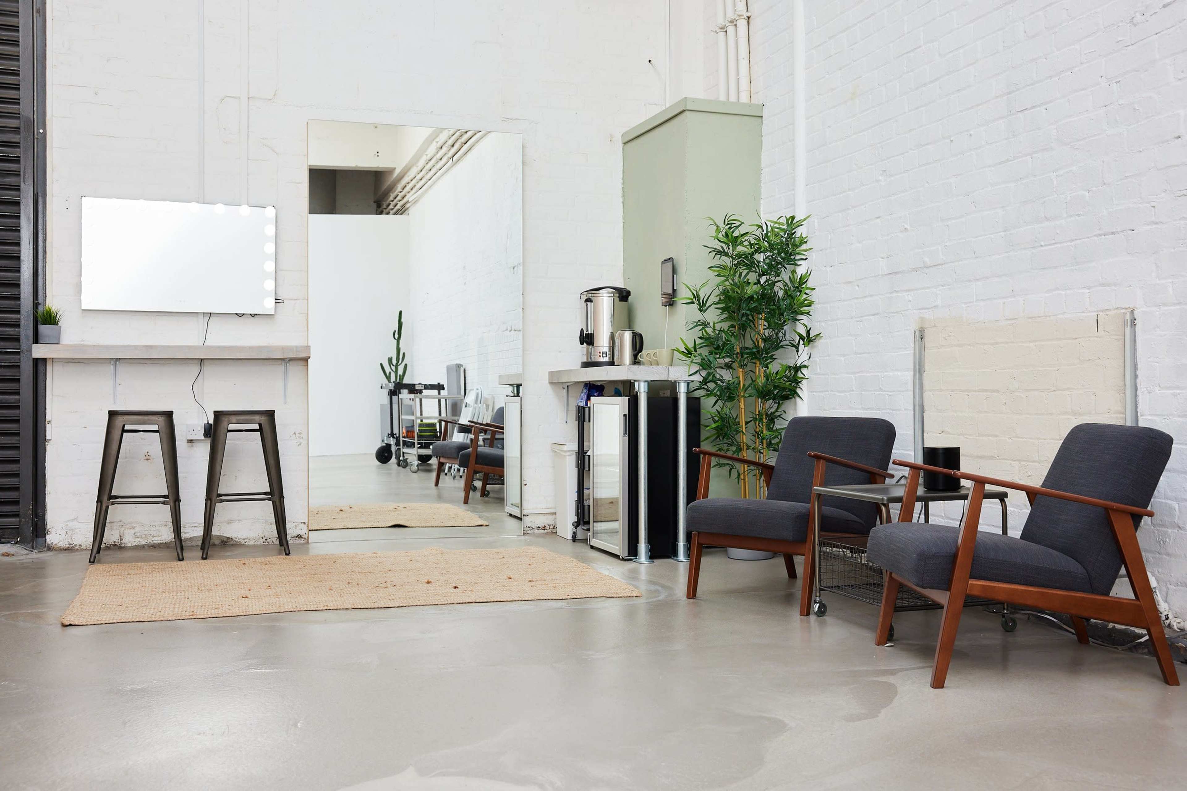 A minimalist interior space with two wooden armchairs, a small table with stools, a mirror, and a plant, all against a backdrop of white brick walls and polished concrete flooring.