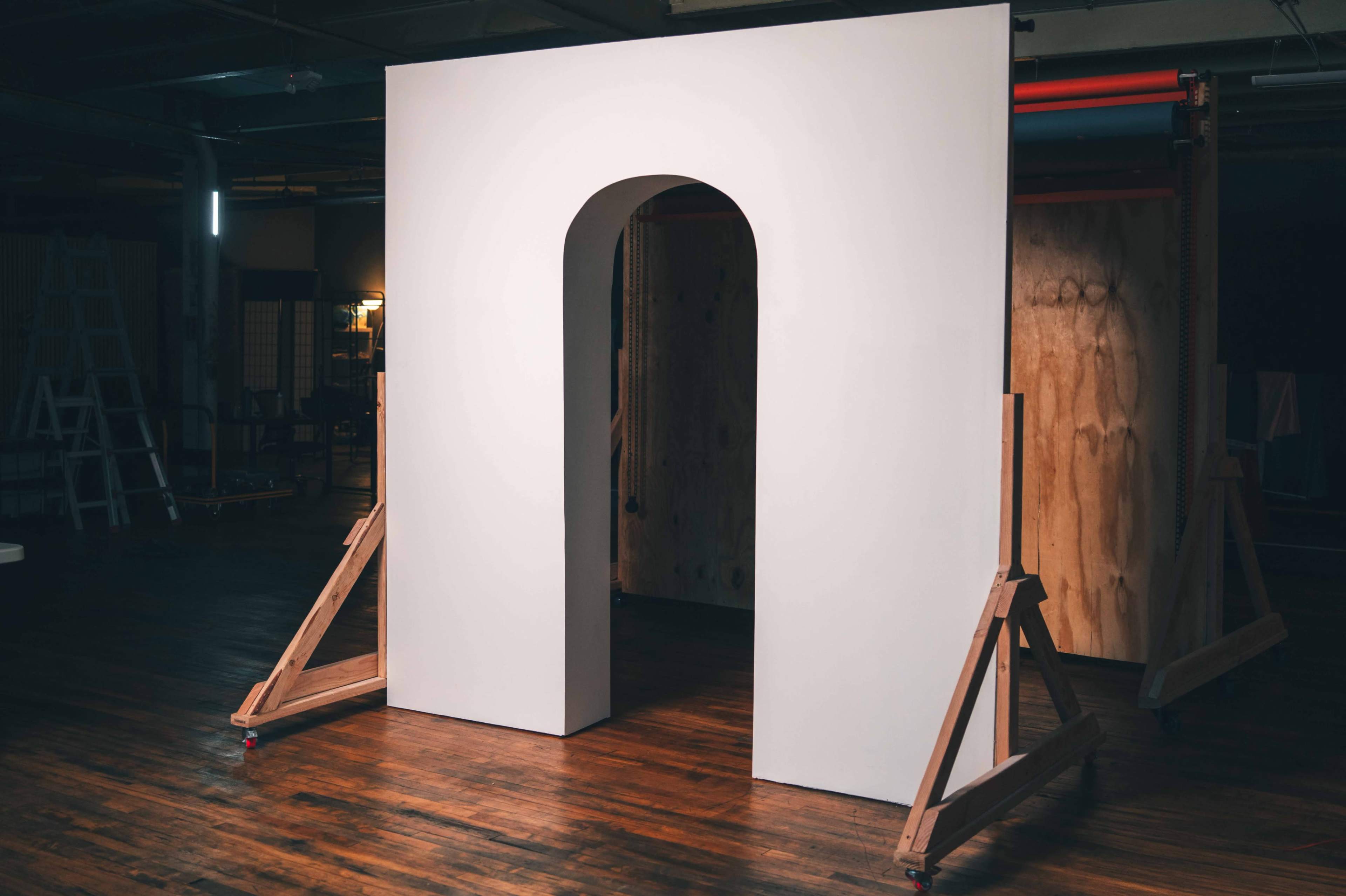 A large, white arch structure stands on a wooden floor, supported by two wooden brackets in a dimly lit room.