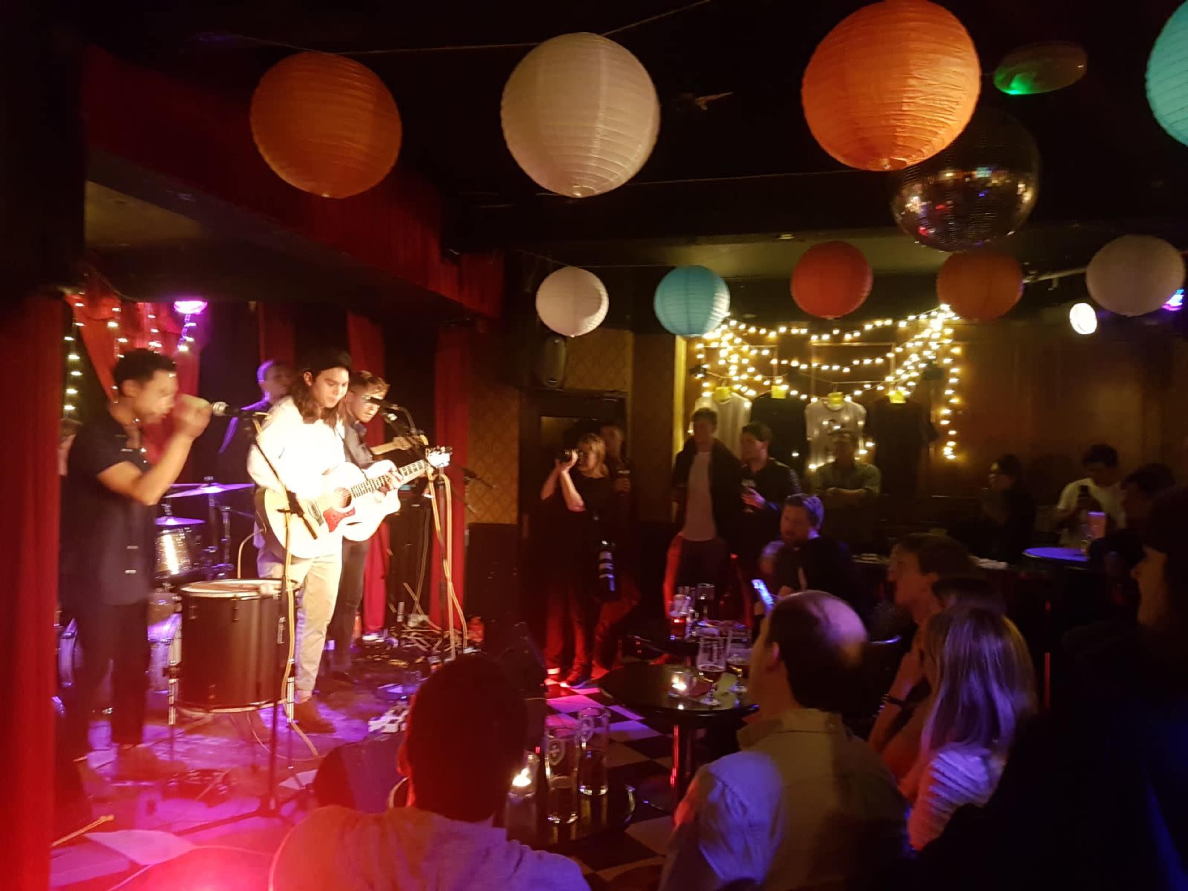 A band performs on stage in a dimly lit bar decorated with colorful lanterns and string lights while an audience watches.