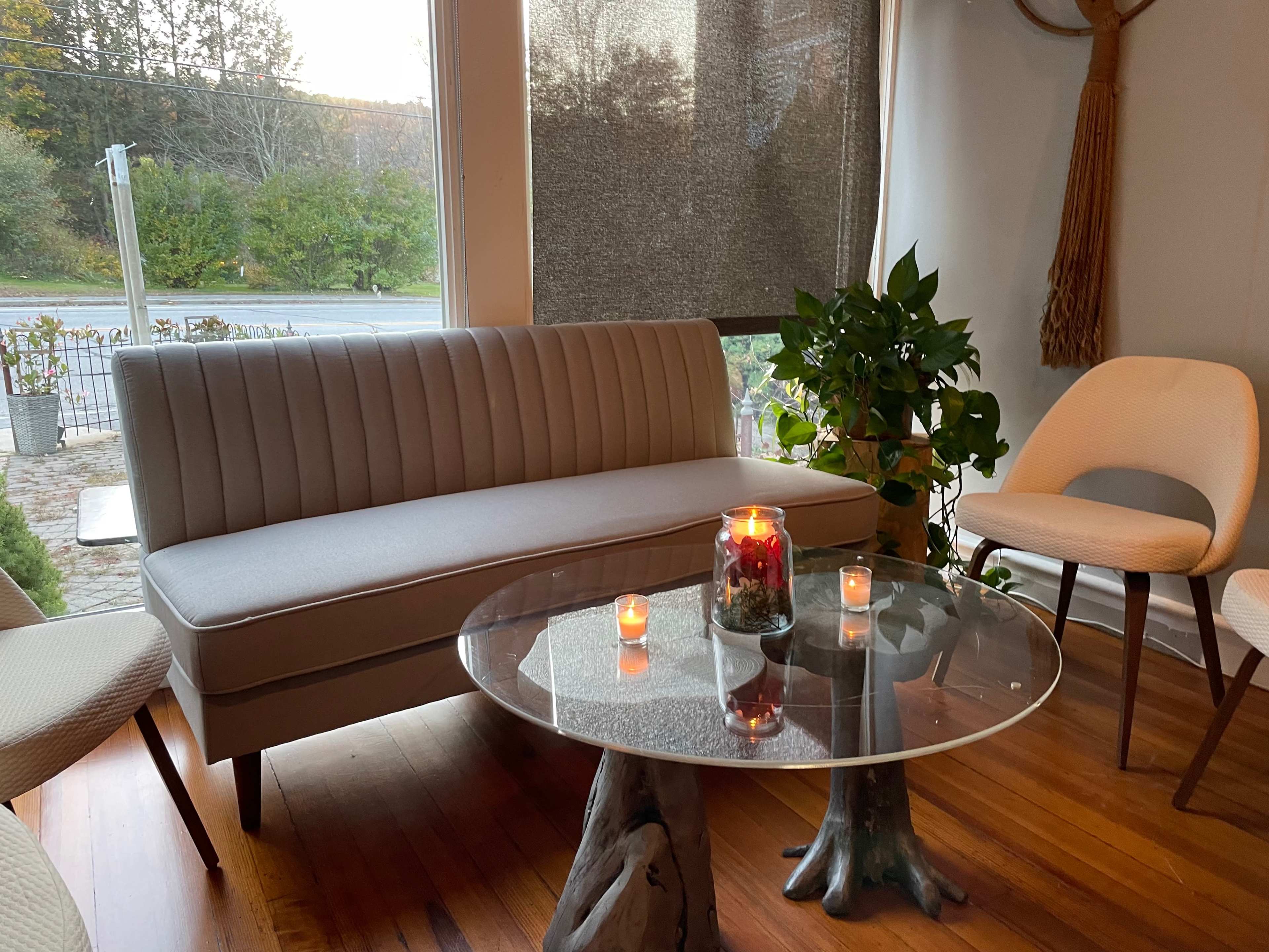 A modern seating area features a light sofa, a glass table with candles, and potted plants beside large windows.